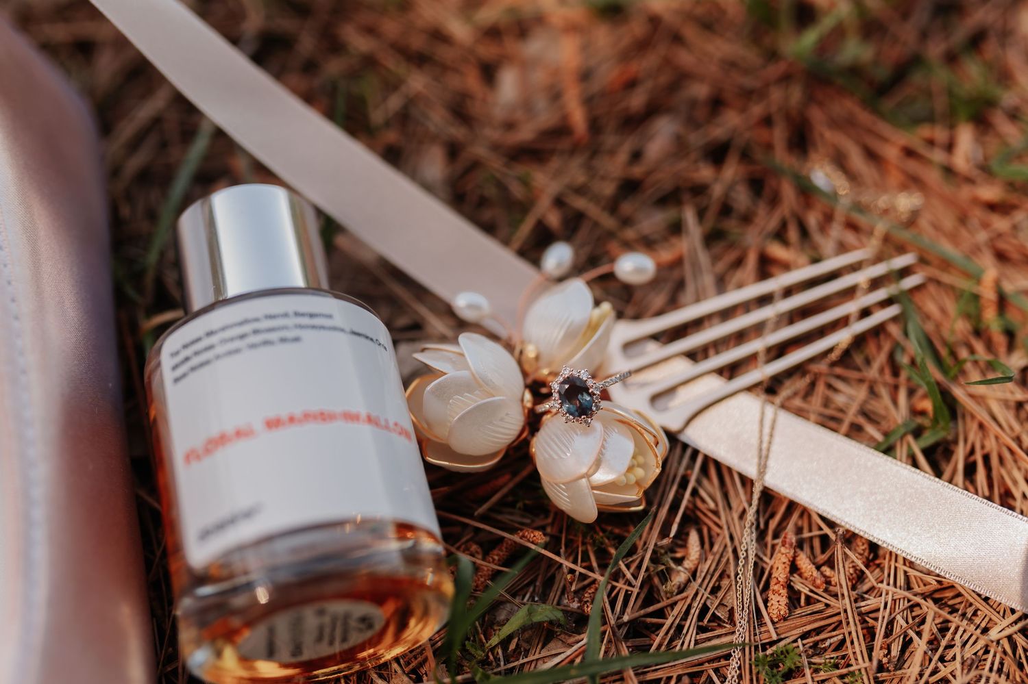 Floral perfume bottle with hair fork and flower accessory on pine needles, elegant wedding flat lay.