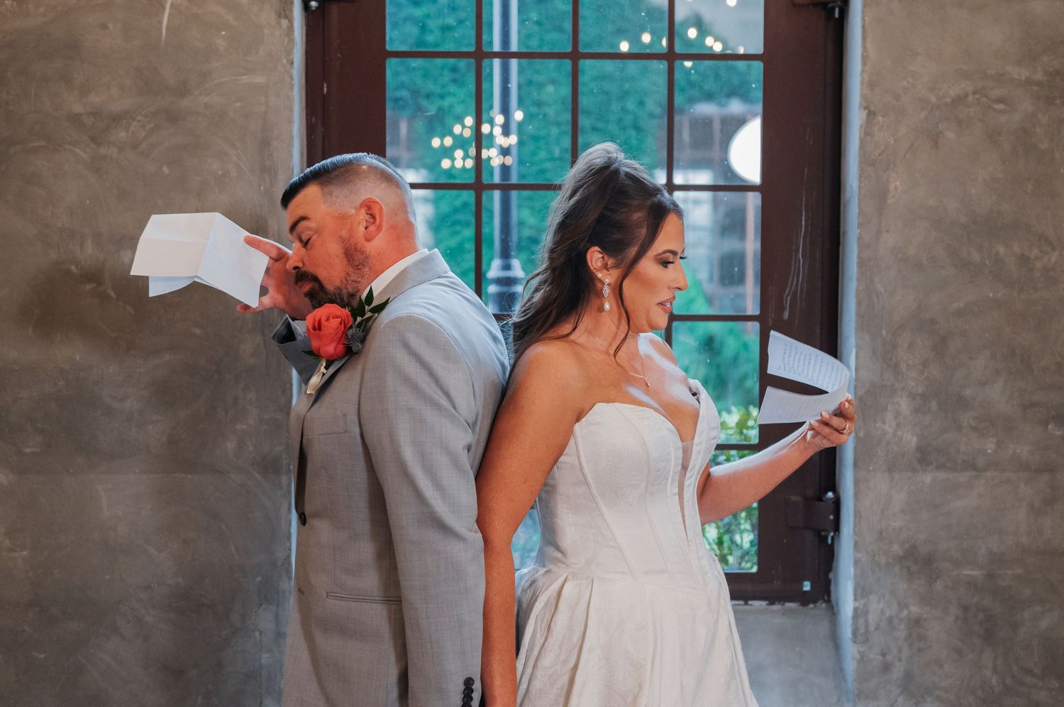 Bride and groom stand back-to-back reading heartfelt vows letters before their wedding ceremony indoors.