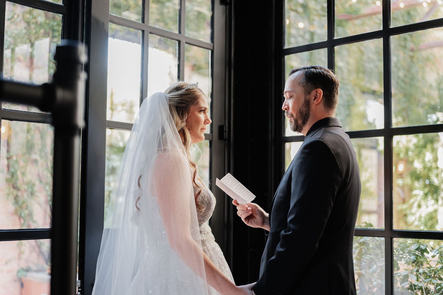 Bride and groom exchanging vows by large black-framed windows, bride in white gown with veil, groom in dark suit holding vows.