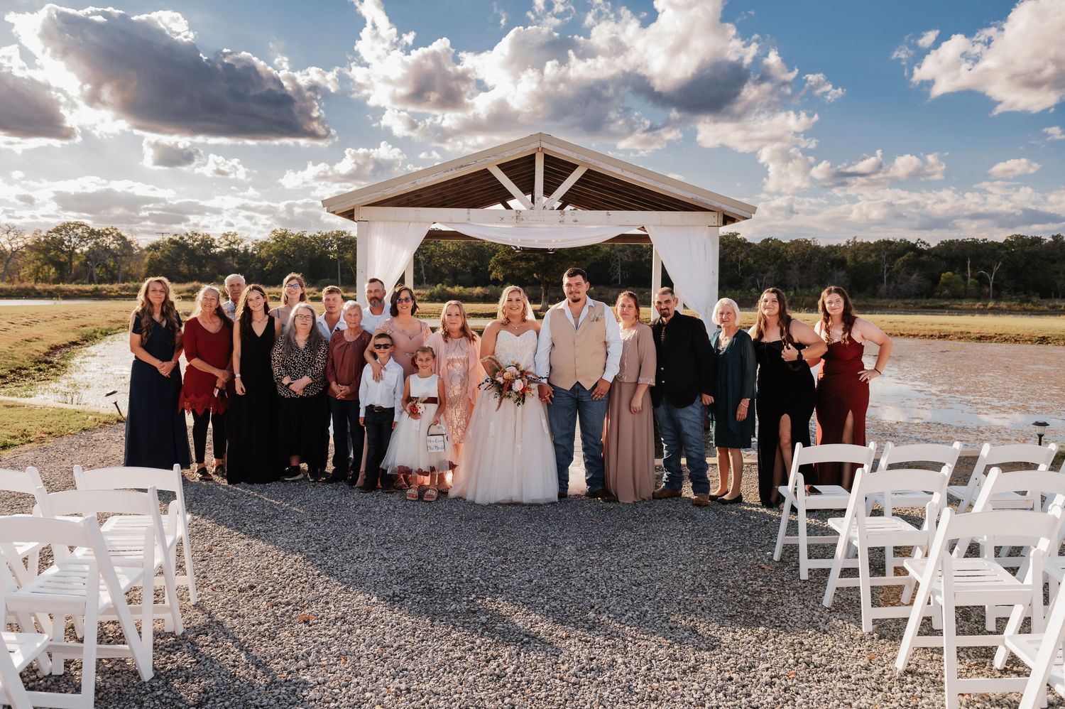 Wedding party poses outdoors near a white pergola on a gravel path, with chairs and a scenic countryside backdrop.