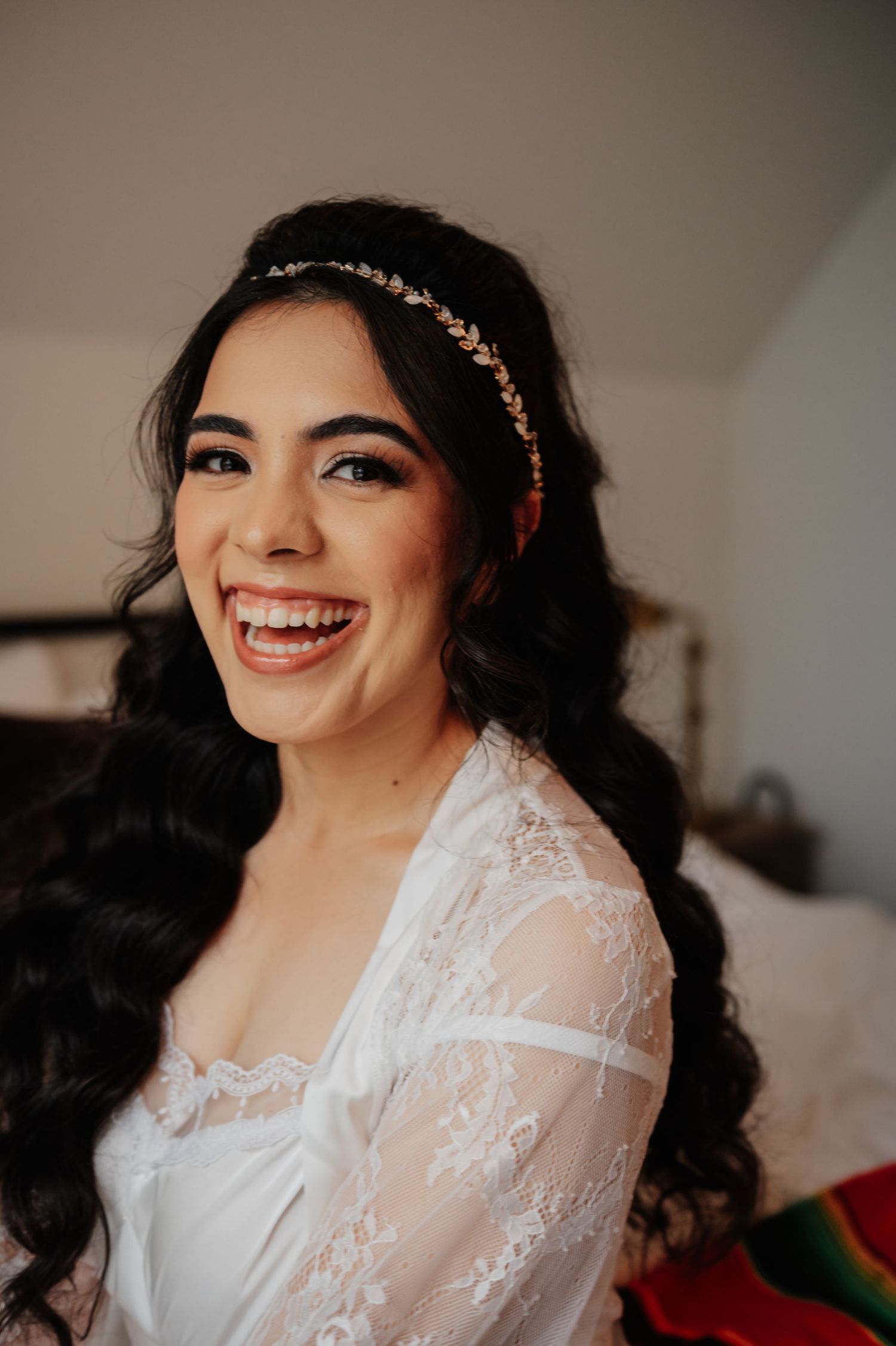 Smiling bride wearing a gold leaf headband and white lace robe with long dark curly hair on her wedding day.