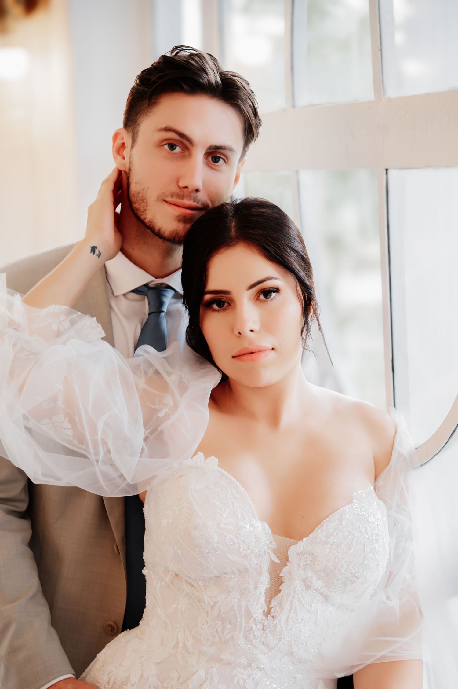 Elegant bride and groom posing together near a bright window, bride in lace off-shoulder gown, groom in gray suit with blue tie.