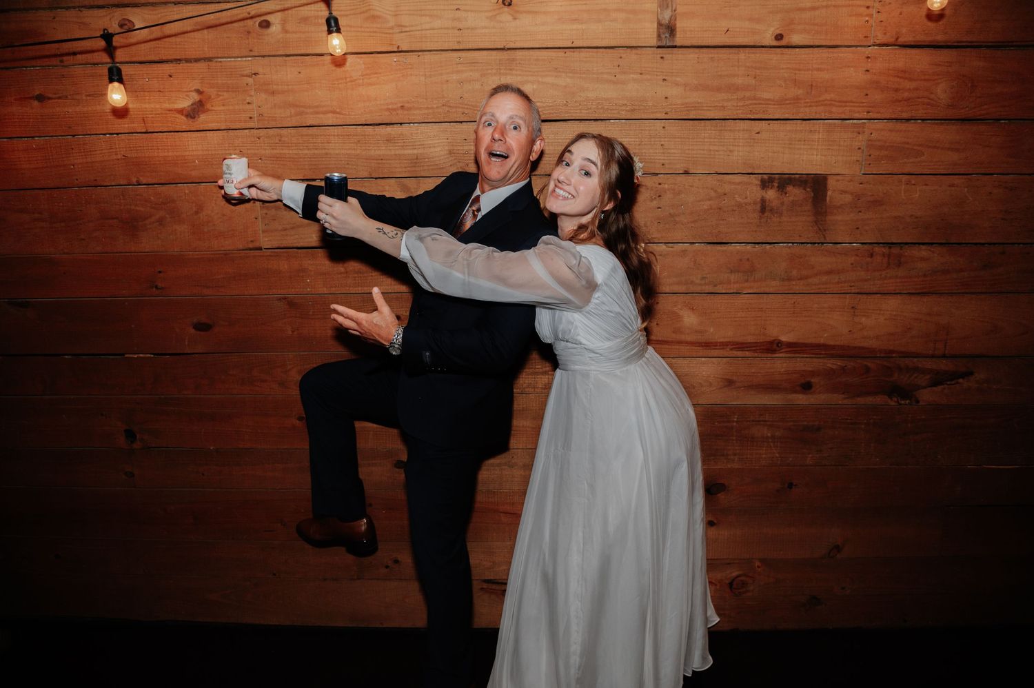 Joyful couple dancing at wedding reception, bride in white gown, groom in suit, rustic wood wall backdrop.