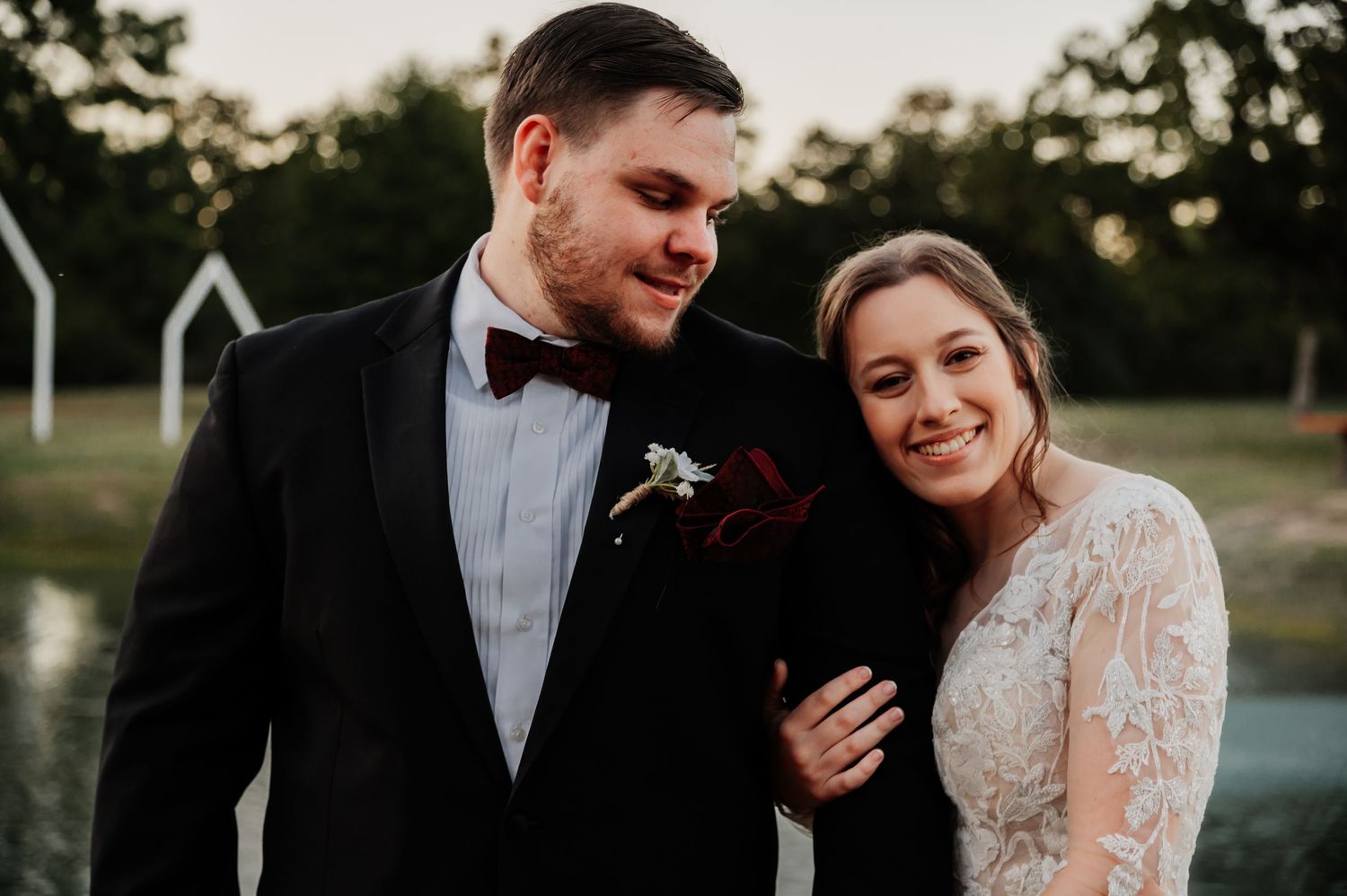 Bride and groom smiling together outdoors at sunset, groom in black tuxedo with boutonniere, bride in lace wedding gown.