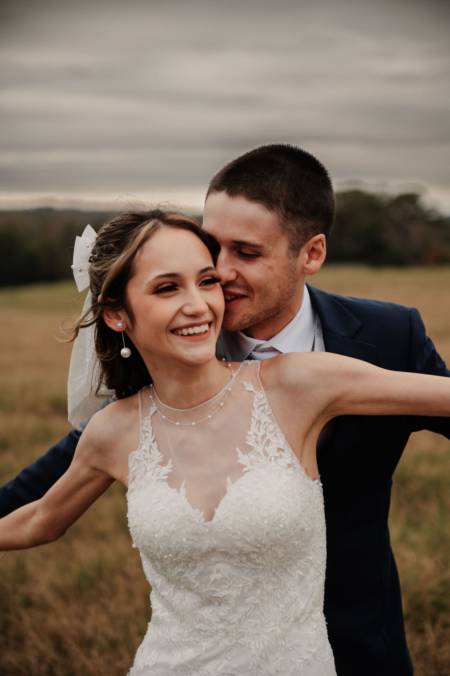 Bride in lace gown laughs joyfully as groom in navy suit kisses her cheek in an open field.