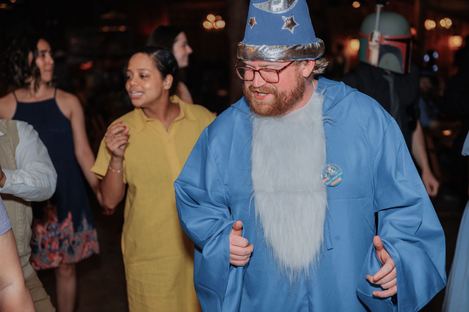 Man in blue wizard costume with fake beard and hat dancing at a party with guests in background.