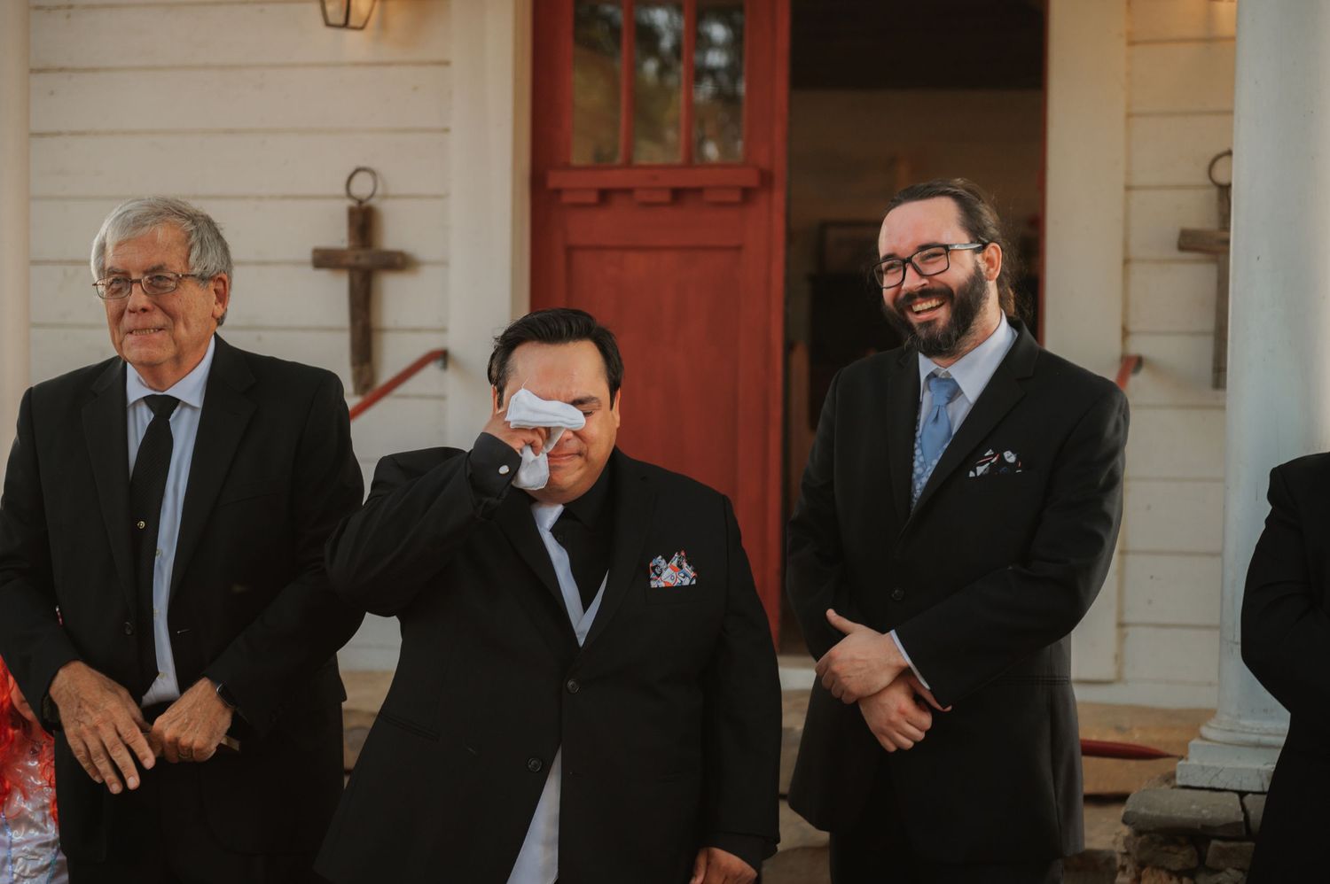 Three men in suits at a wedding, one emotional wiping tears, another smiling warmly near a red door.