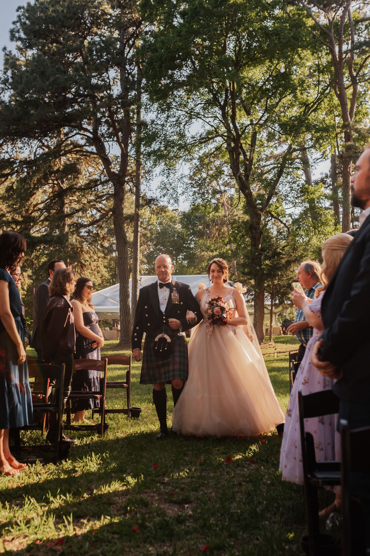 Bride and groom walk down outdoor aisle at sunset wedding ceremony, groom wearing traditional Scottish kilt.