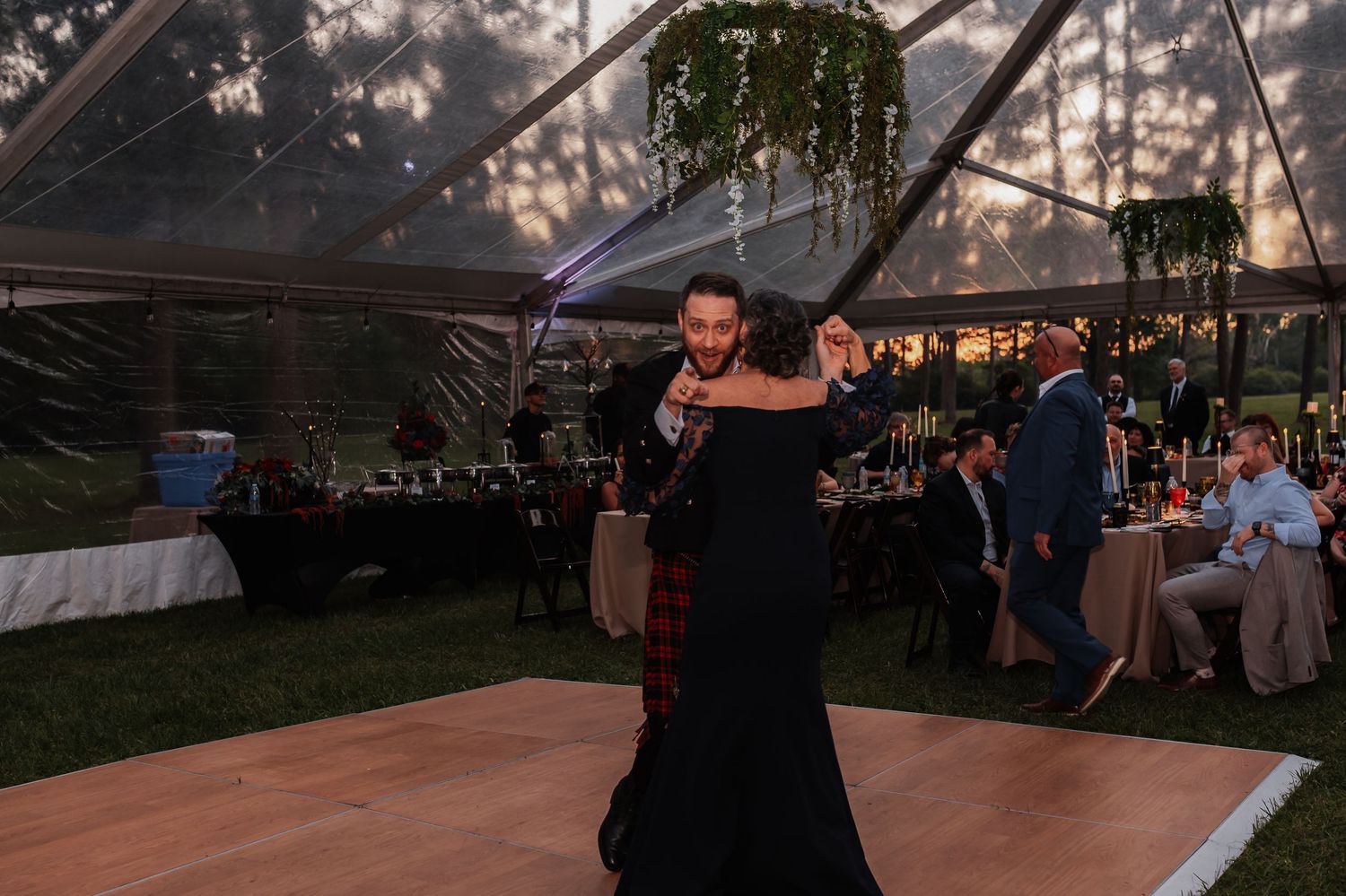 Couple sharing a romantic first dance under a clear tent at an outdoor wedding reception at dusk.