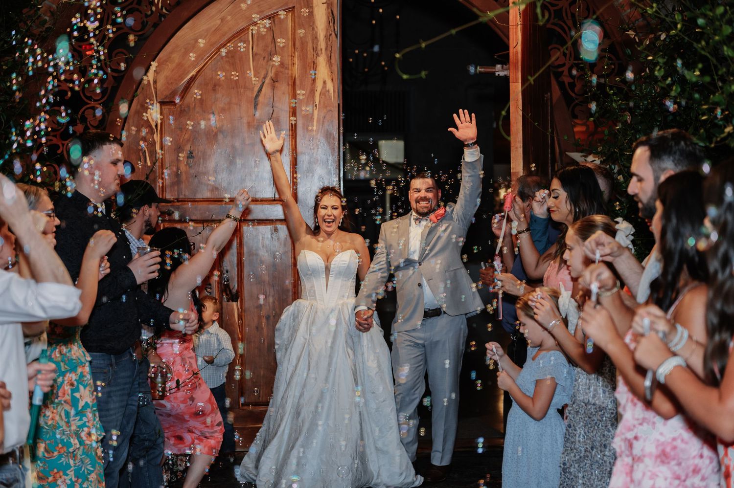 Bride and groom exit wedding venue celebrating with guests throwing confetti near rustic wooden doors.
