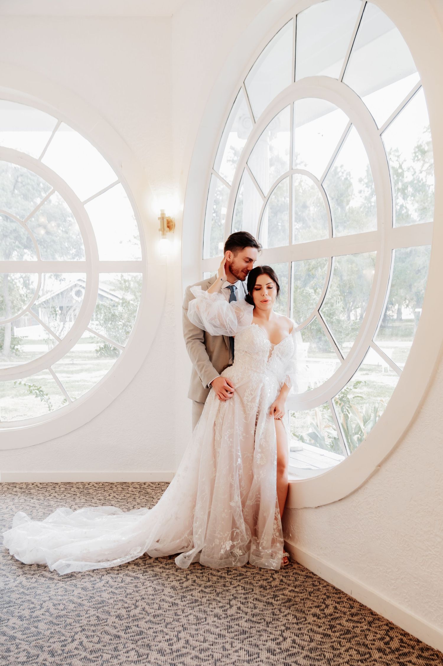 Bride in lace gown with train and groom in suit pose before circular windows in elegant white venue.
