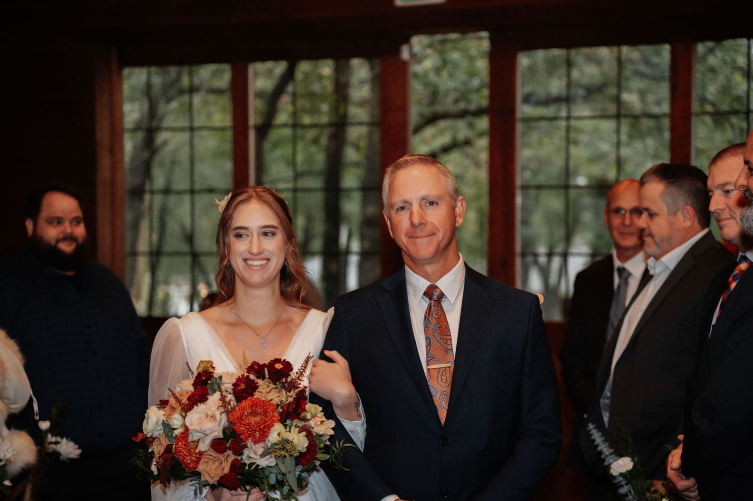 Bride holding bouquet walks down aisle with escort at indoor wedding ceremony.