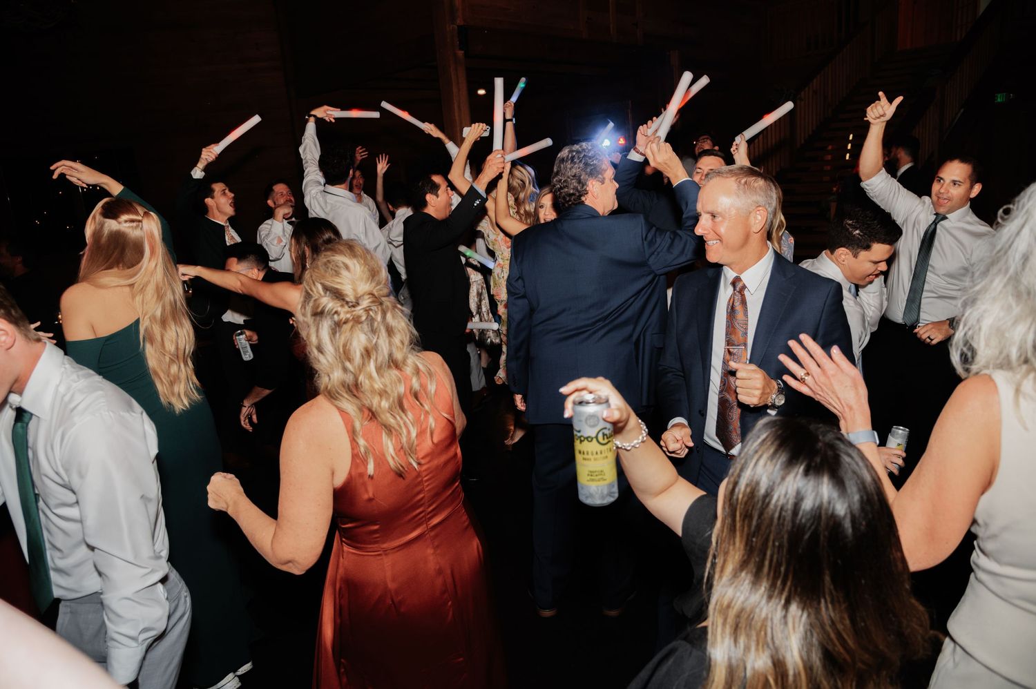 Guests dancing and celebrating at a lively wedding reception with glow sticks inside a dimly lit venue.