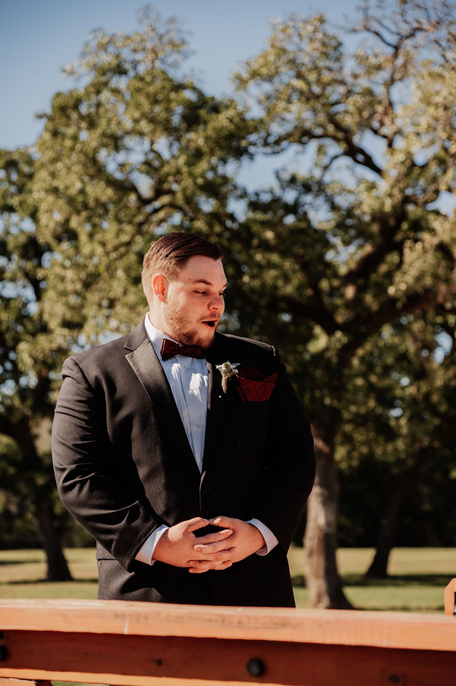 Groom in black tuxedo with bow tie and boutonniere stands outdoors at wedding ceremony among lush green trees.