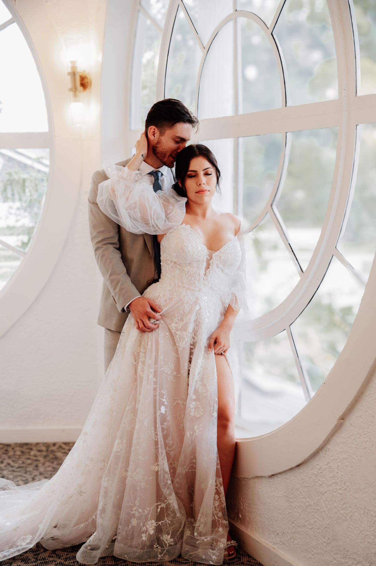 Bride and groom pose romantically by an arched window, bride in a floral lace gown with off-shoulder sleeves.