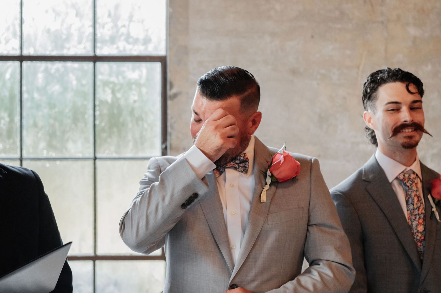 Emotional groom wiping tears in gray suit with pink boutonniere and bow tie, standing beside smiling groomsman at wedding ceremony.