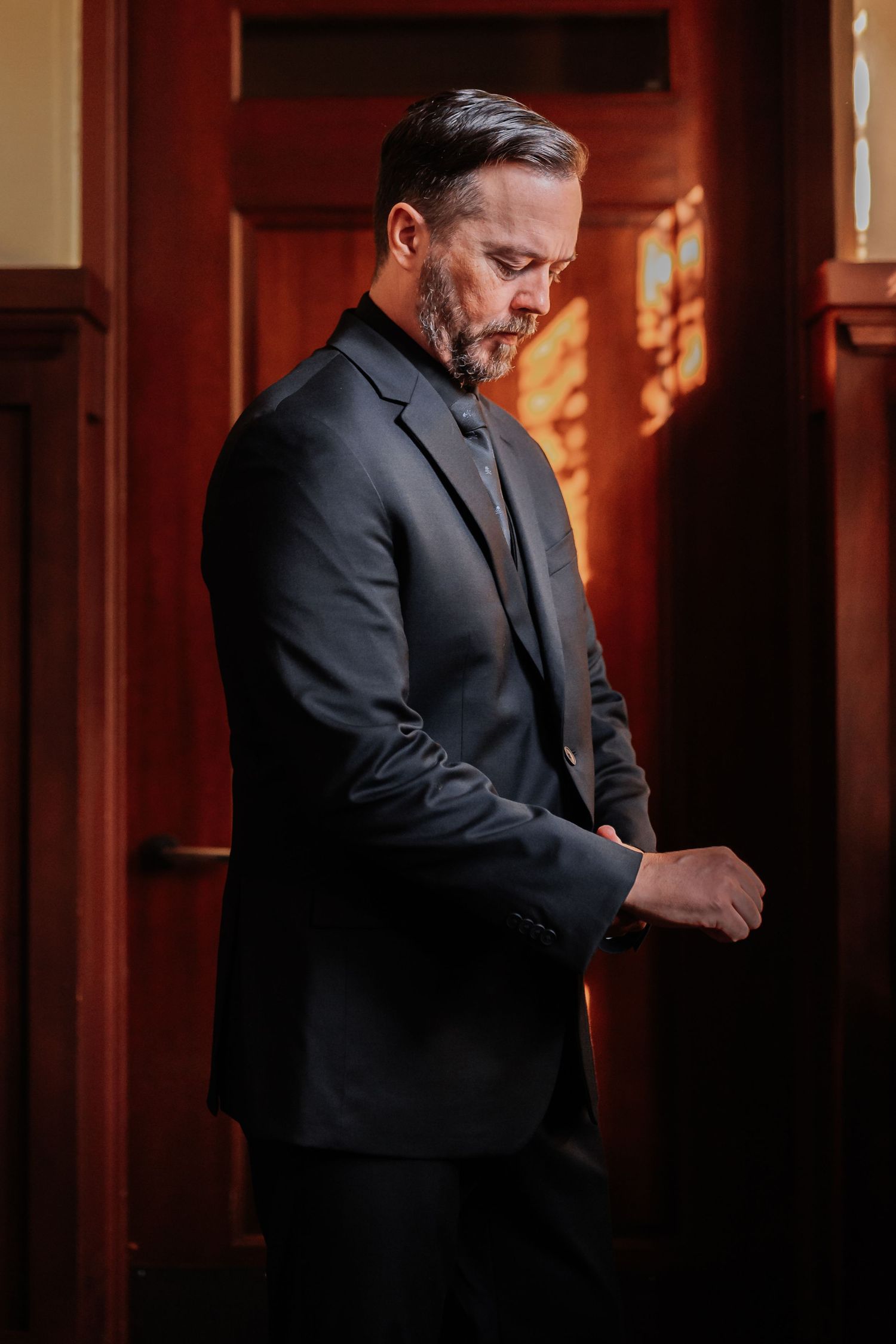 A middle-aged man in a dark suit adjusts his cufflinks by wooden doors with warm golden light.