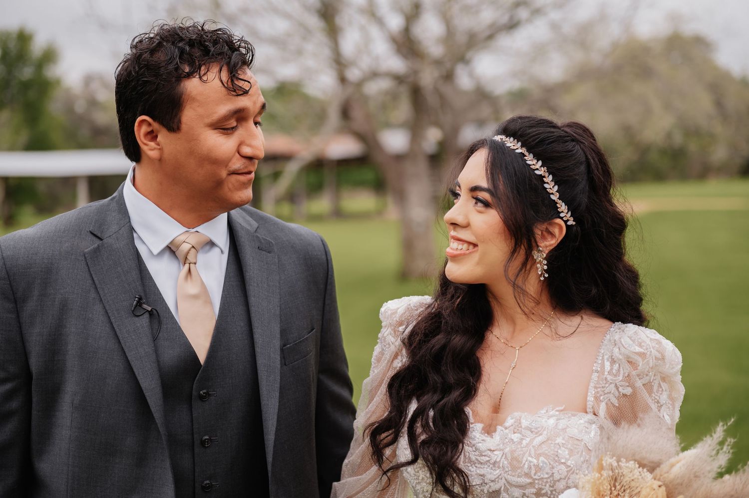 A bride and groom share a romantic glance outdoors, she in a lace gown with floral headpiece, he in a gray suit.