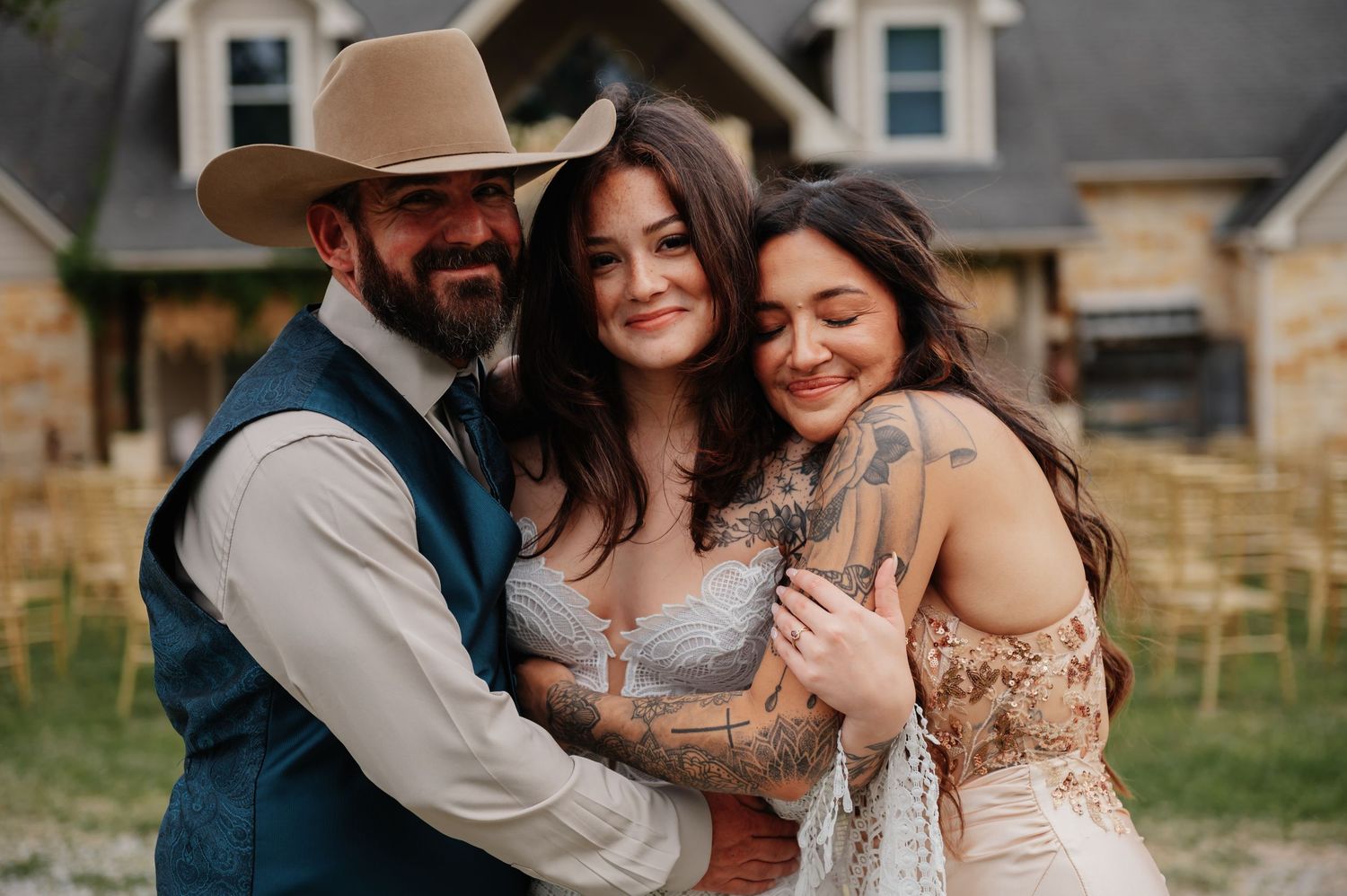 A groom in a cowboy hat and vest embraces two women in wedding attire outdoors at a rustic venue.