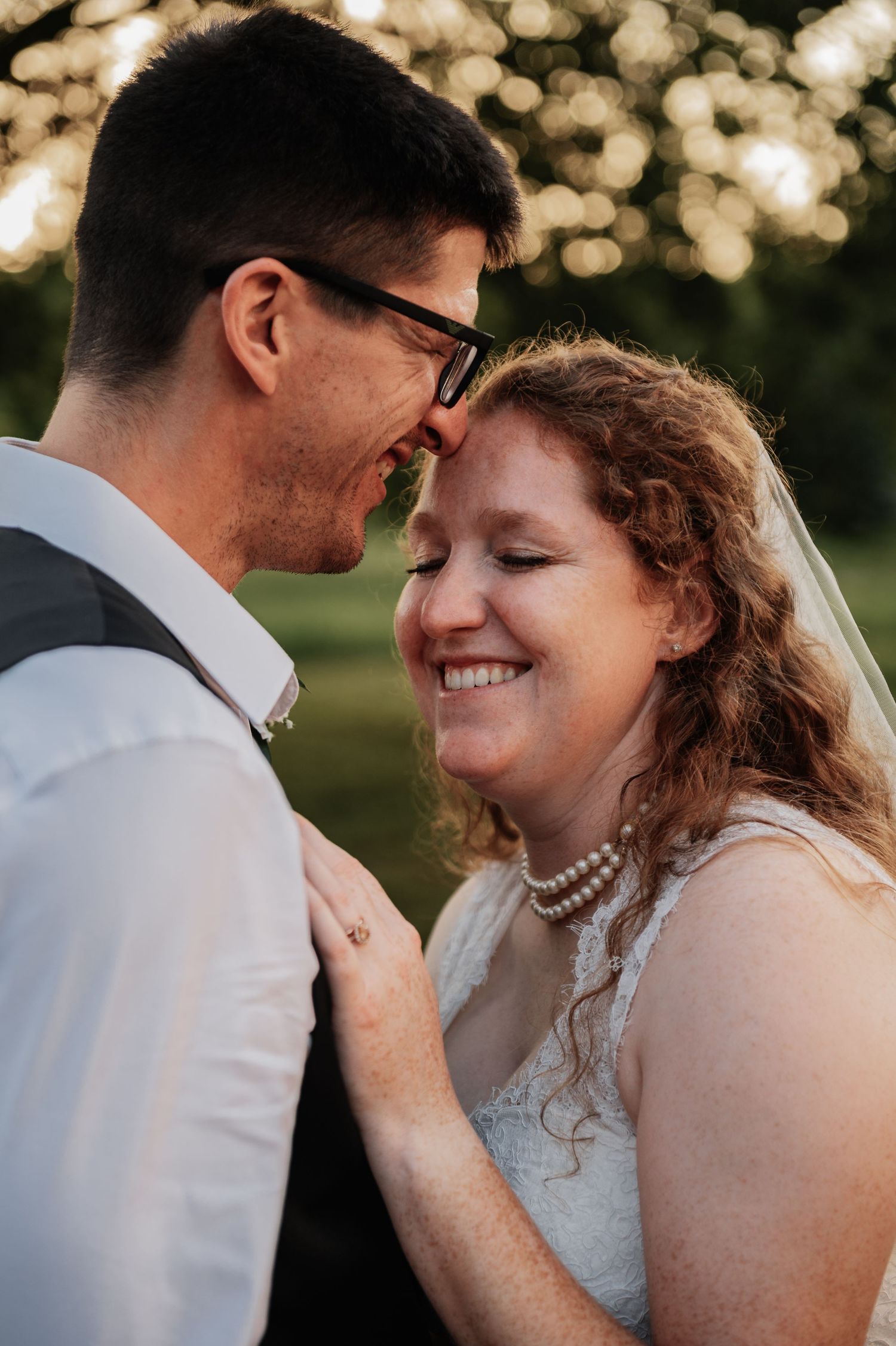 A smiling bride in lace dress and pearl necklace touches foreheads with groom in glasses at golden hour outdoor wedding.
