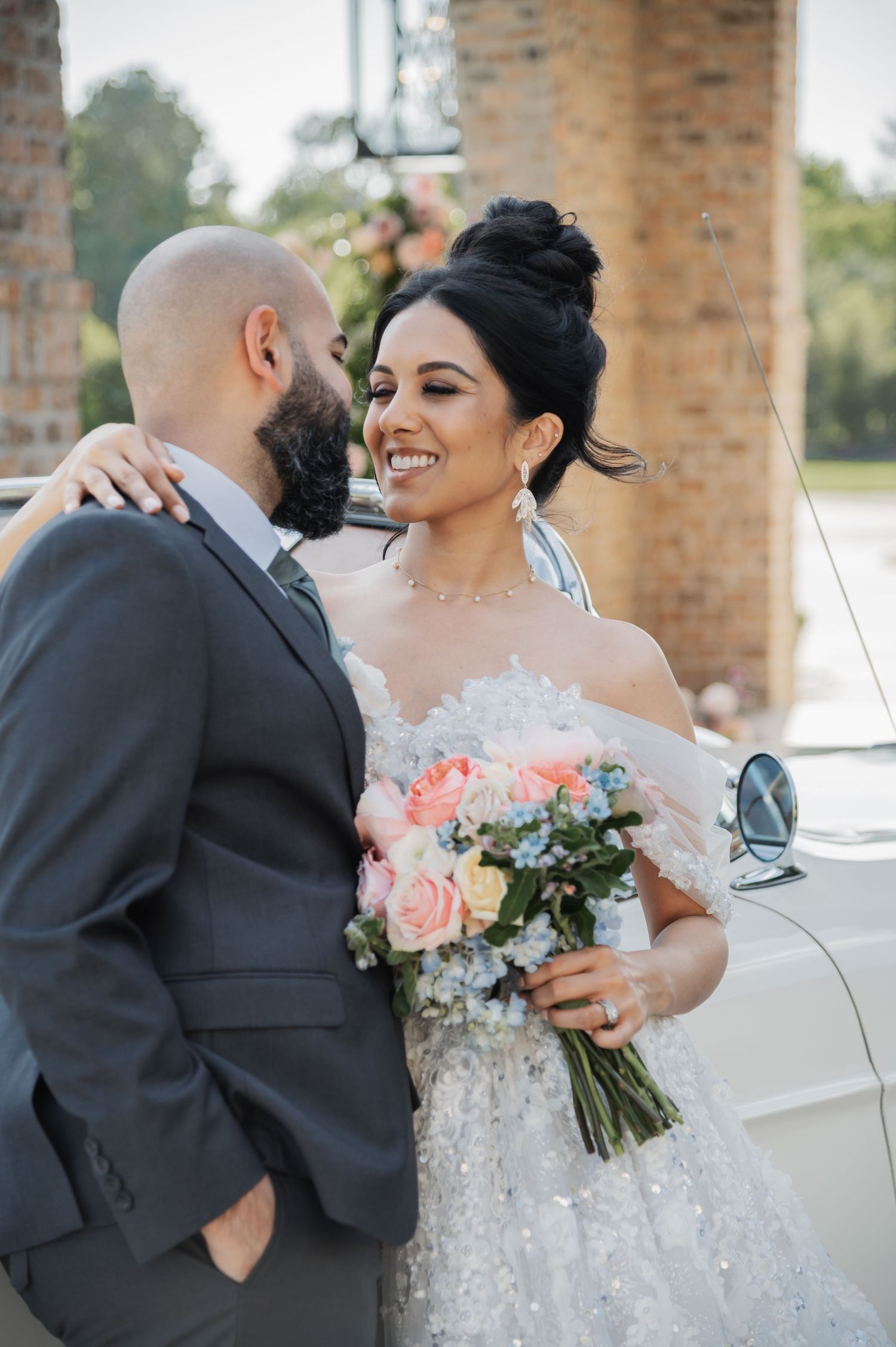 Smiling bride holds a pastel bouquet while embracing her groom near a vintage white car at their wedding.