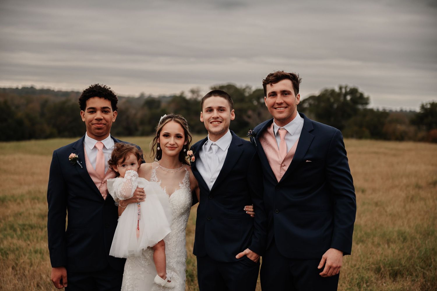 Wedding party poses in a field, bride in white gown with groom and groomsmen in navy suits with pink ties.