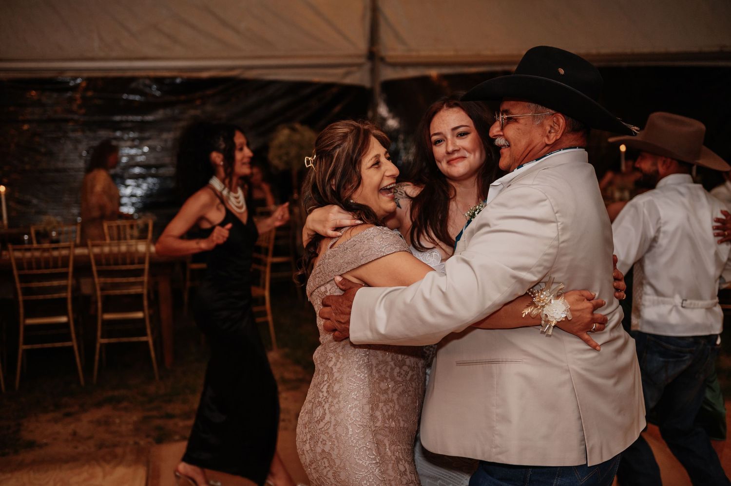 Three guests share a warm group hug on the dance floor at an elegant outdoor wedding reception.