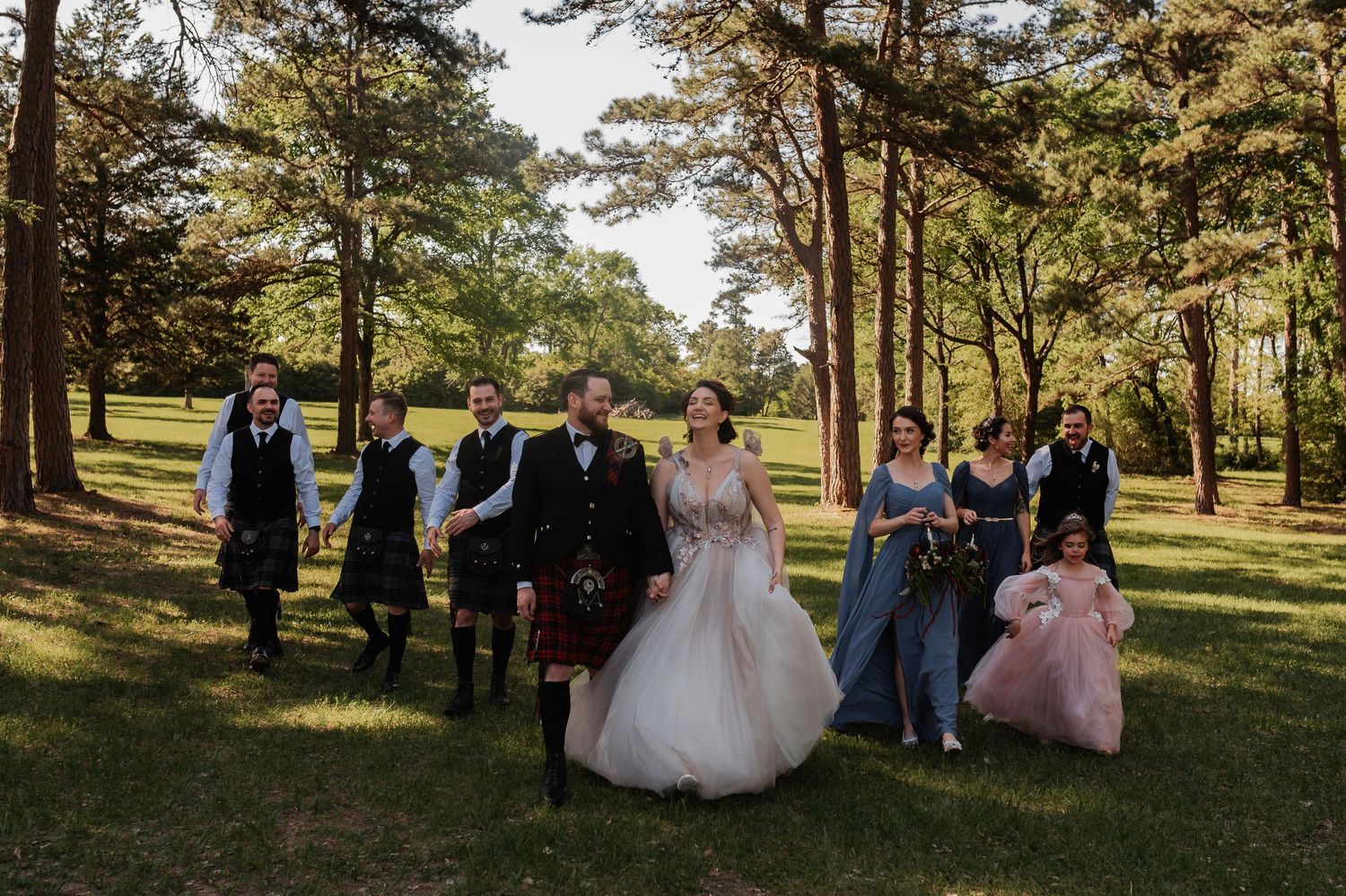 Wedding party walks through lush green forest, bride in white gown with groom in Scottish kilt surrounded by bridesmaids.