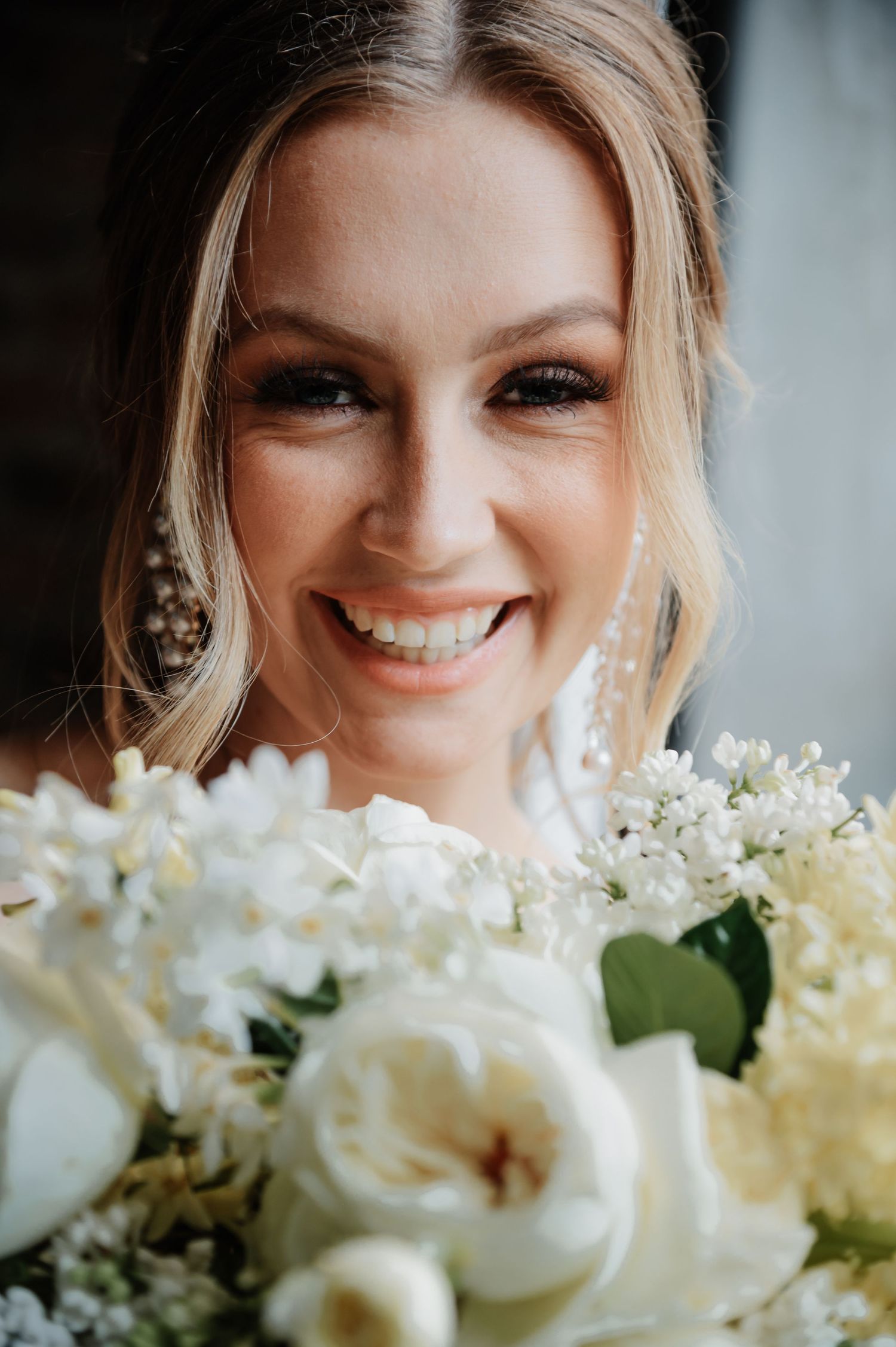 Radiant bride with pearl drop earrings smiling behind lush white rose and lilac wedding bouquet.