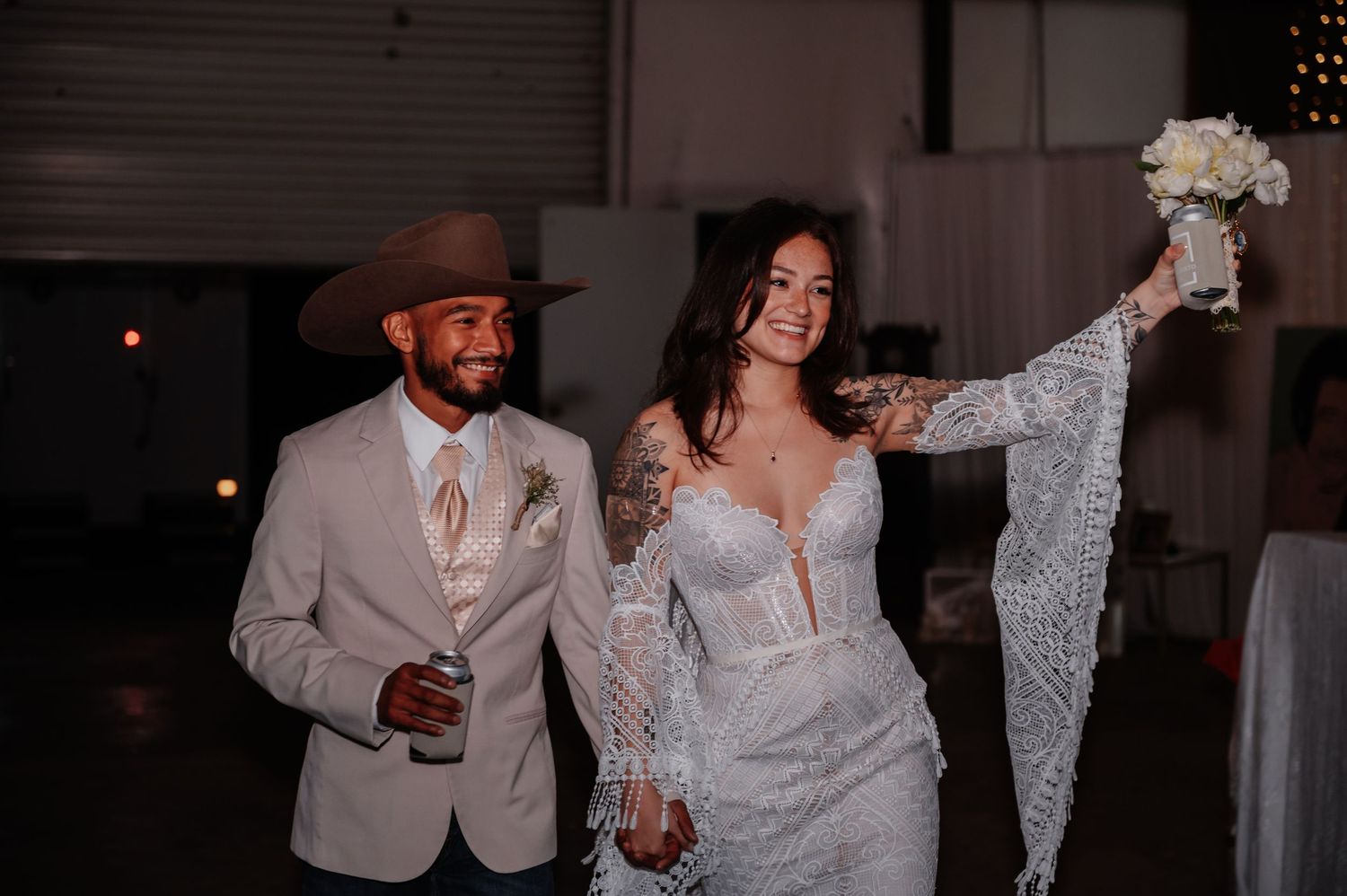 Newlyweds walking triumphantly at their wedding reception, groom in tan suit and cowboy hat.