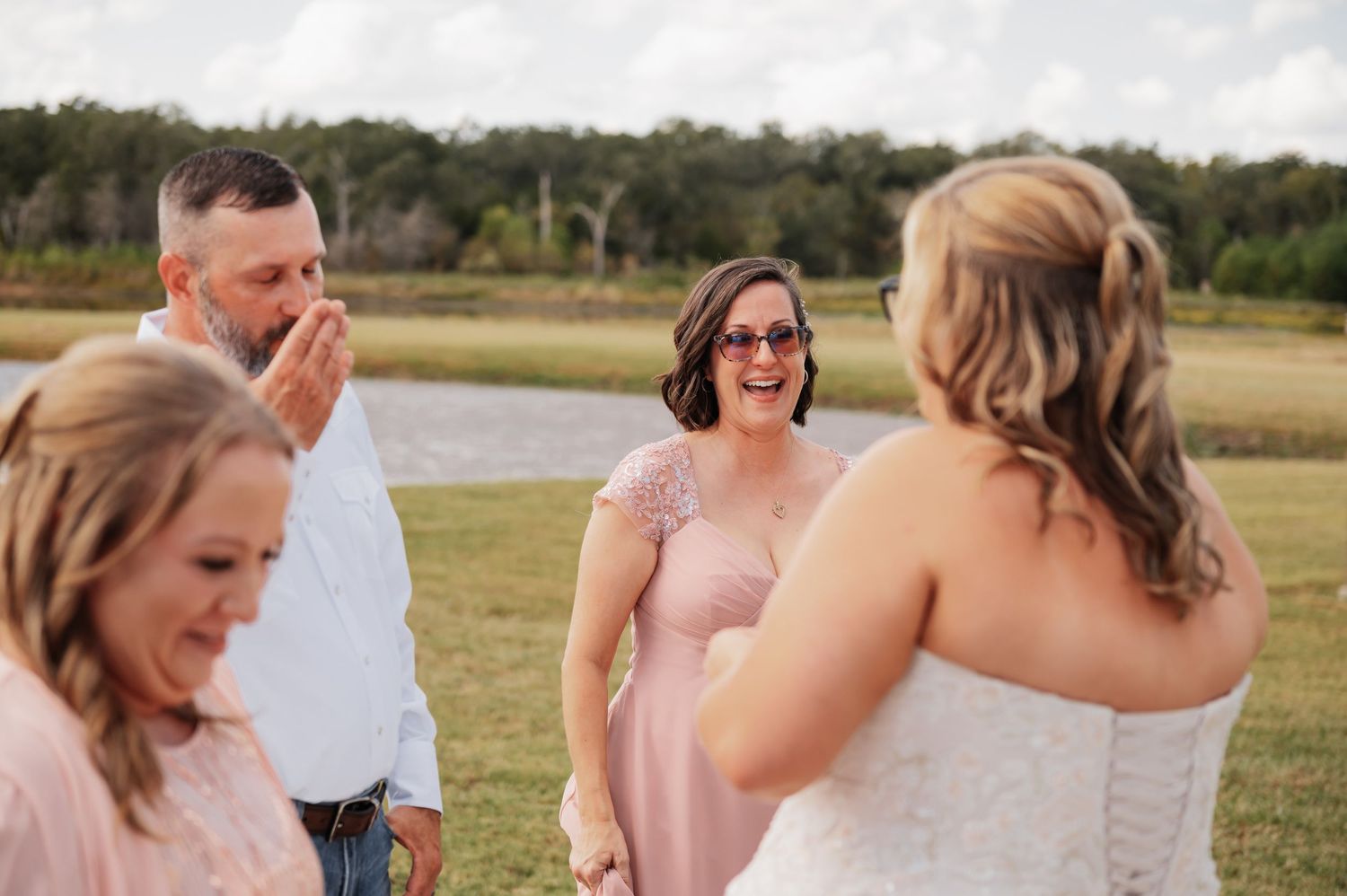 Emotional wedding moment with groom covering mouth in tears, surrounded by bridesmaids in pink dresses outdoors.