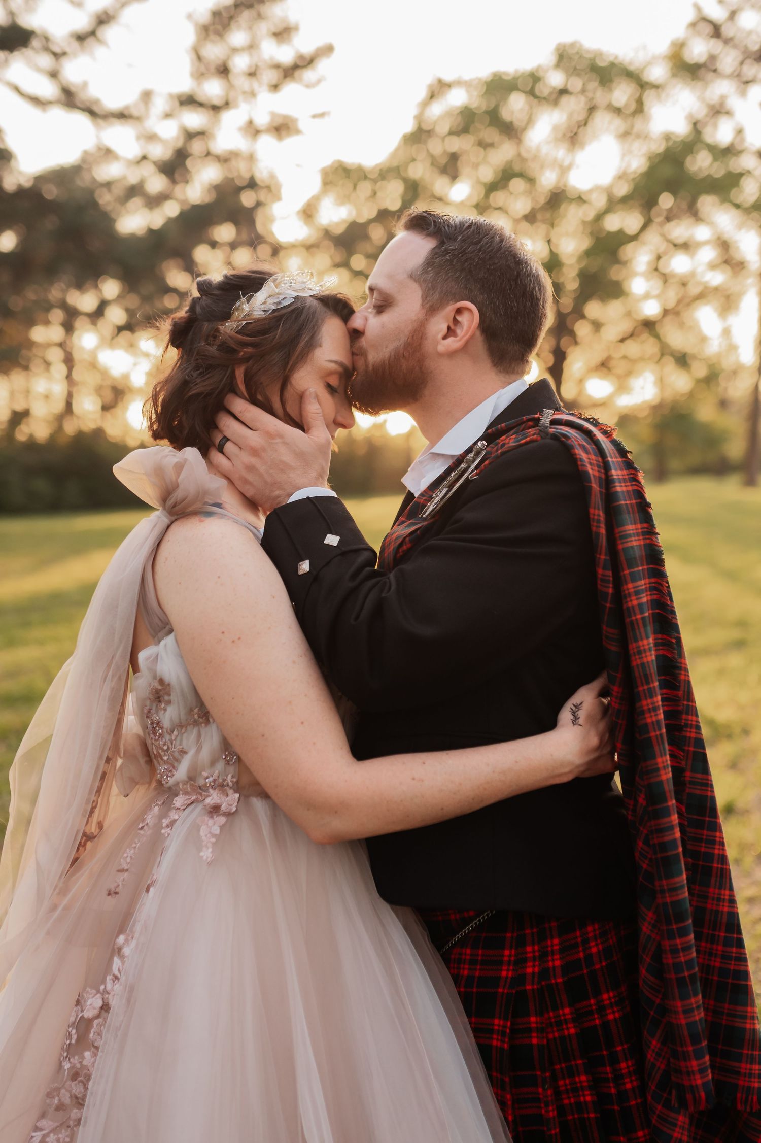 Bride and groom share a romantic kiss at golden hour, groom wearing a red tartan sash over his suit.