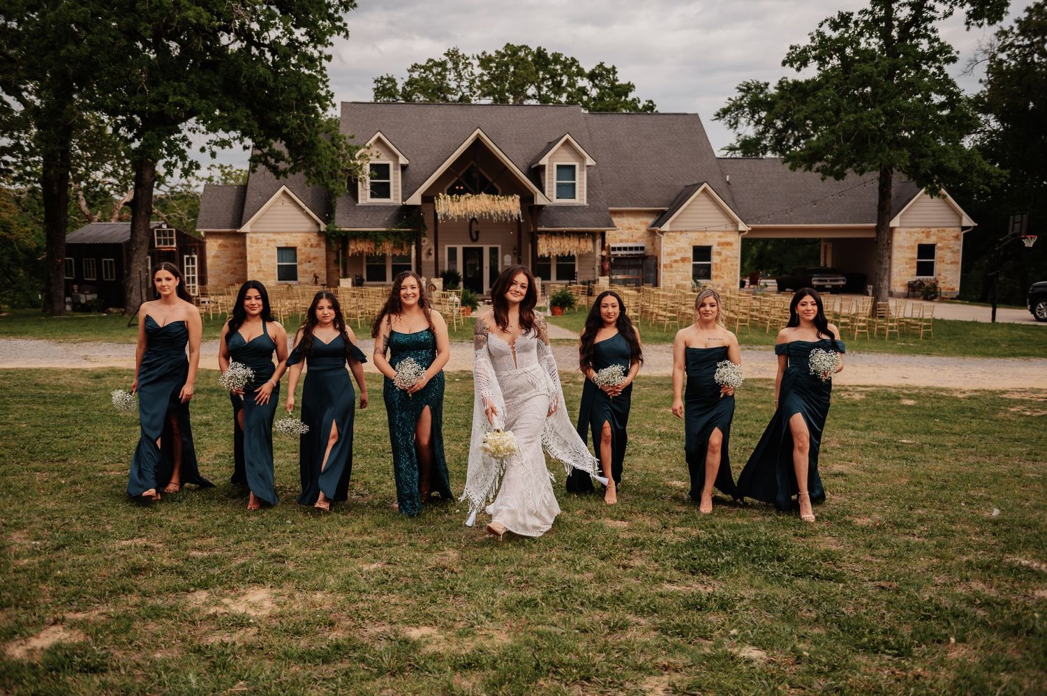 Bride and bridesmaids in elegant teal gowns walk together laughing outside a beautiful rustic farmhouse.