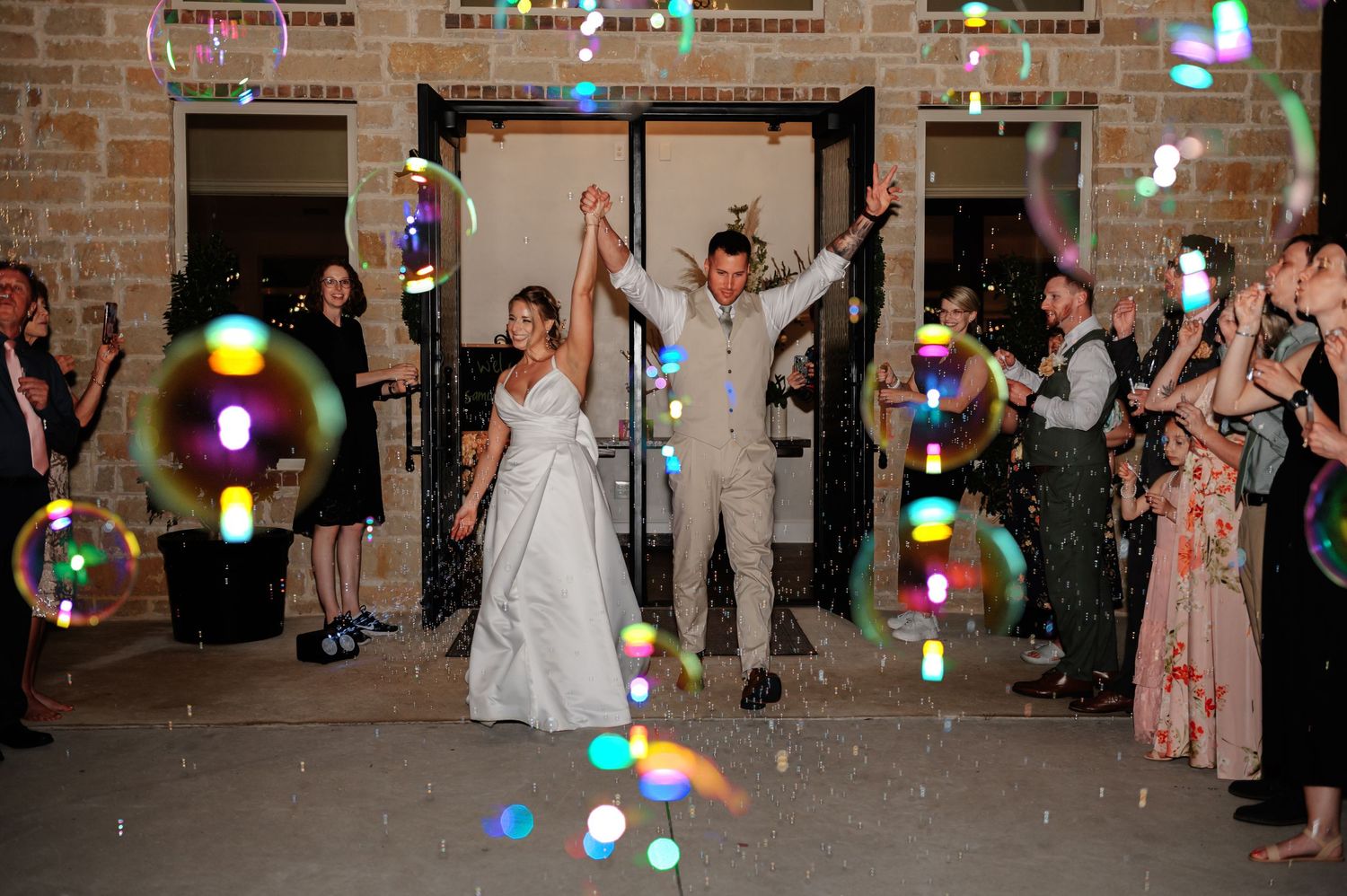 Bride and groom celebrate wedding exit surrounded by colorful floating bubbles while guests cheer outside stone venue.