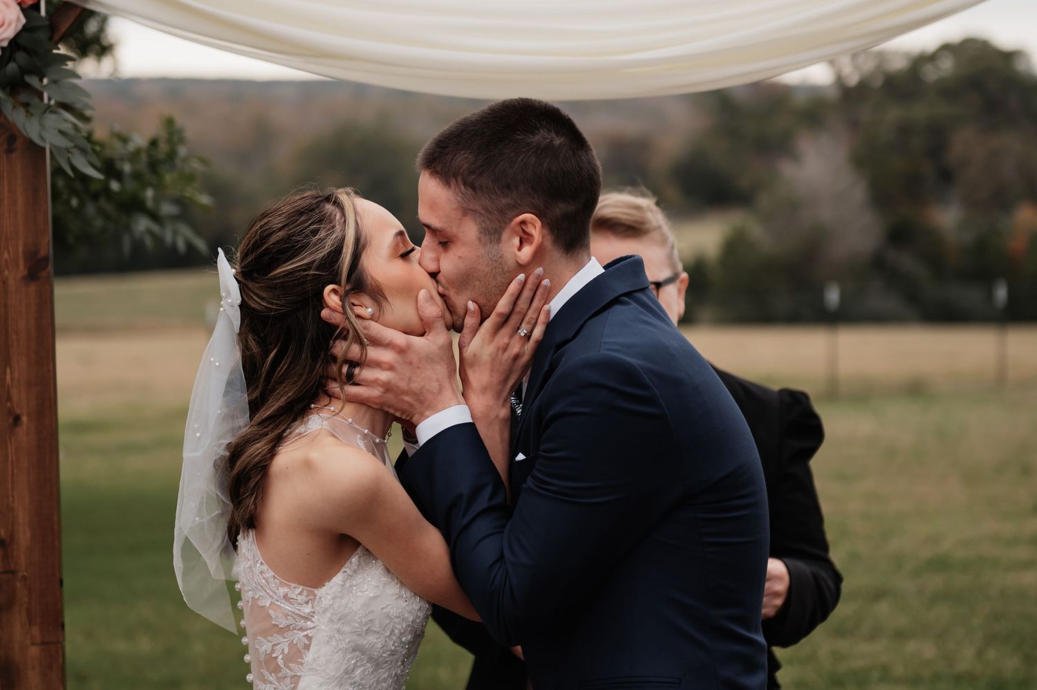 Bride and groom share first kiss at outdoor wedding ceremony with rustic arch backdrop.