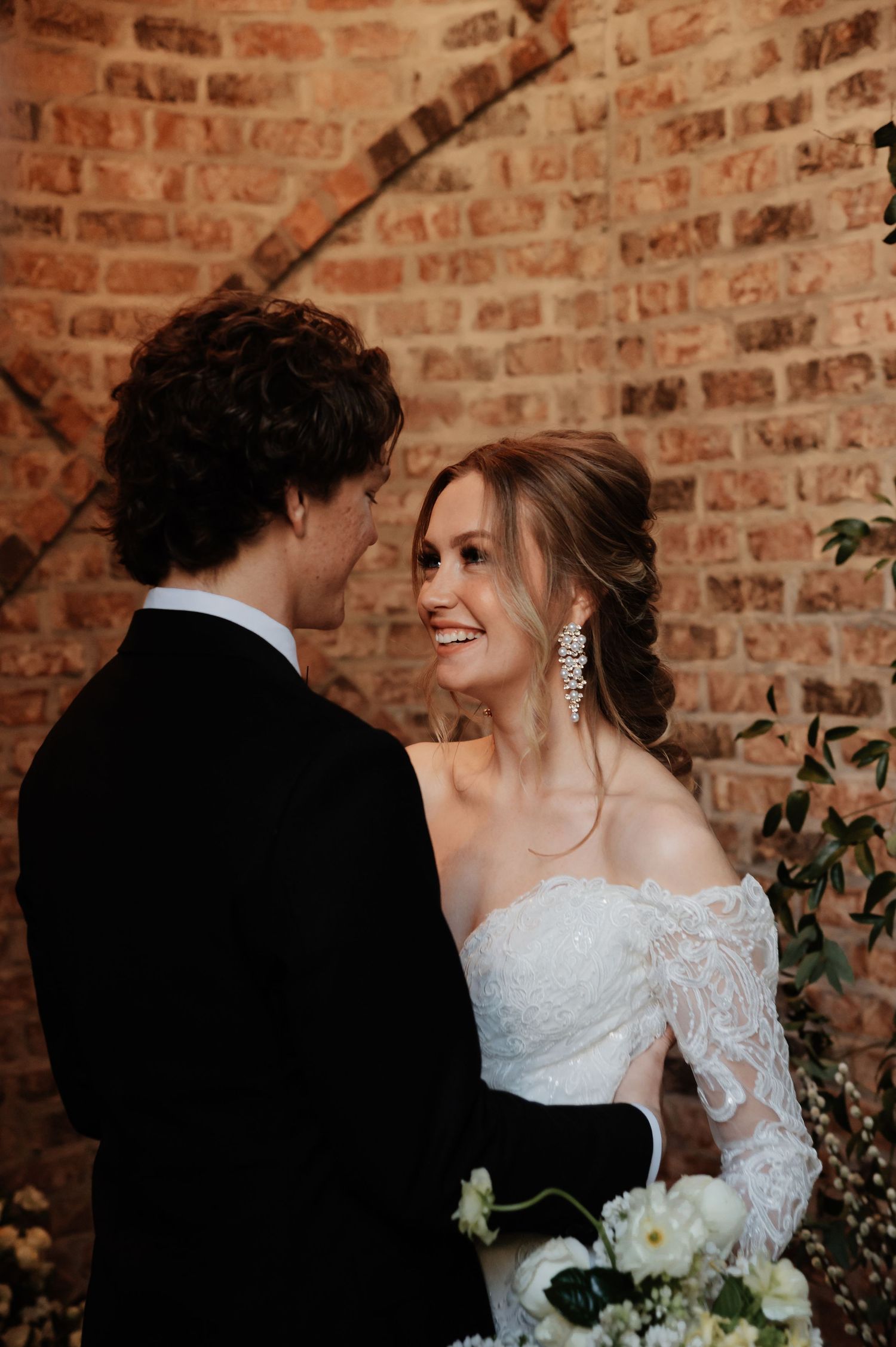 Bride in off-shoulder lace gown with crystal earrings smiling at groom against rustic brick wall backdrop.
