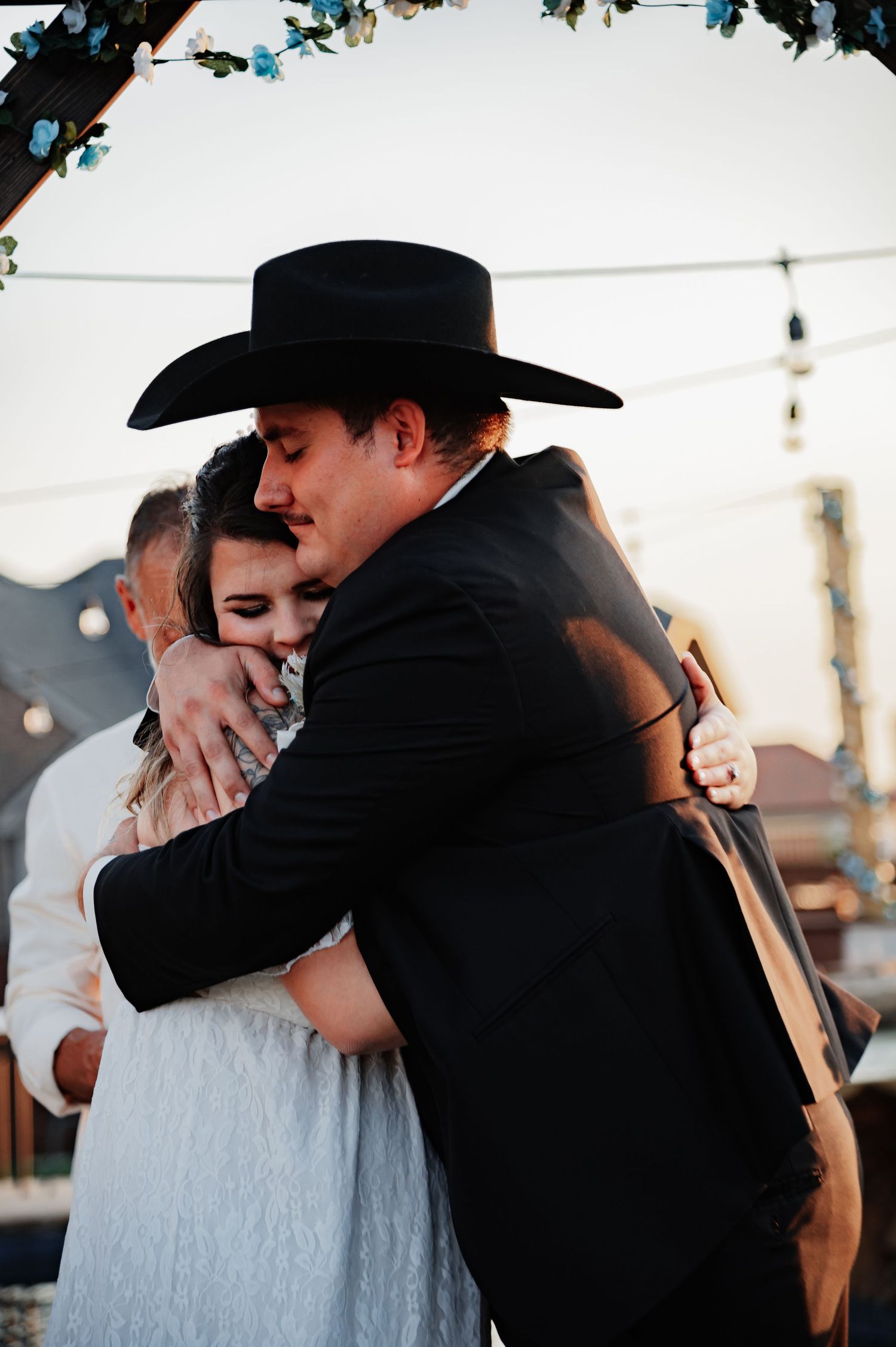 A couple shares an emotional embrace at their western-themed wedding, groom wearing a black cowboy hat and suit.