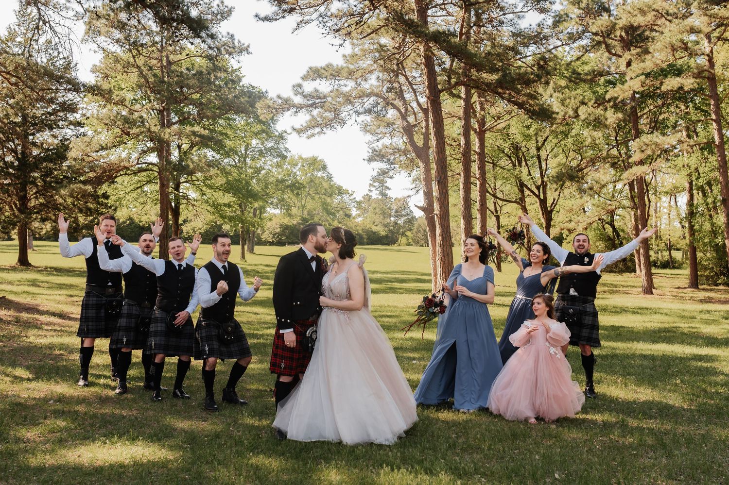 Wedding party celebrates outdoors as bride and groom kiss, groomsmen in kilts and bridesmaids in blue gowns among pine trees.