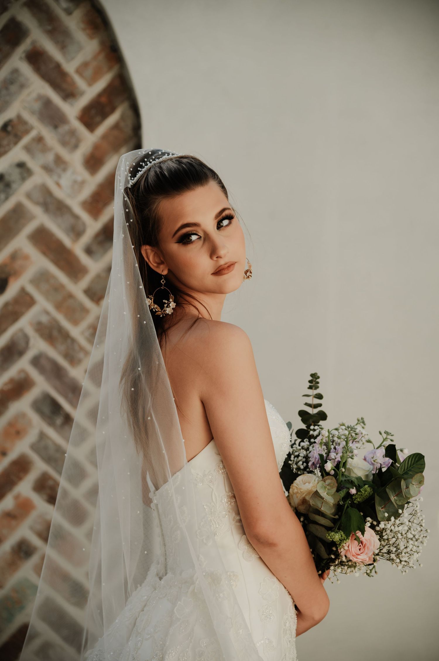 Elegant bride in a strapless gown with veil and tiara poses against a rustic brick wall holding flowers.