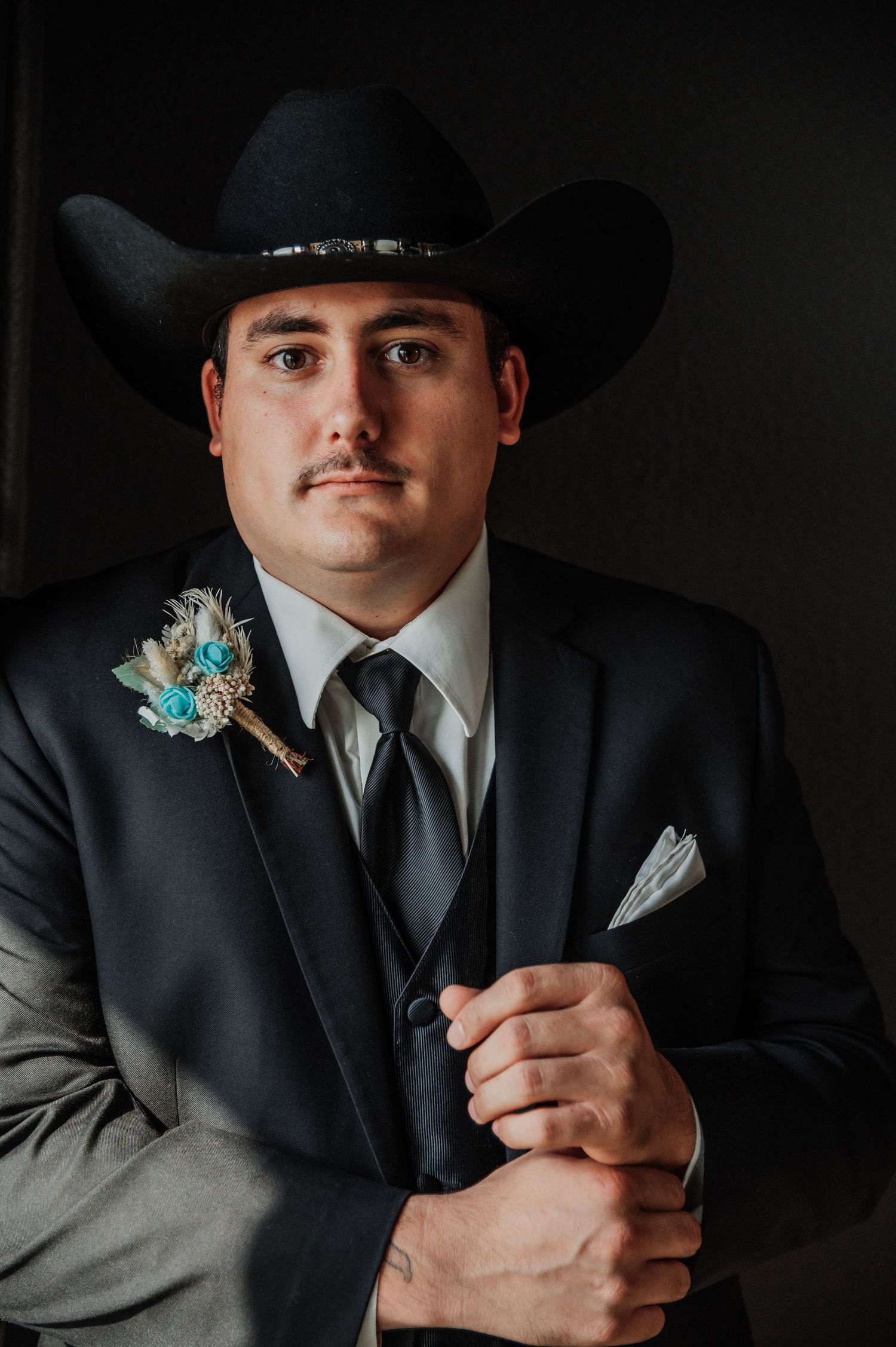 A groom in a black cowboy hat and dark suit with a teal floral boutonniere poses confidently for a wedding portrait.