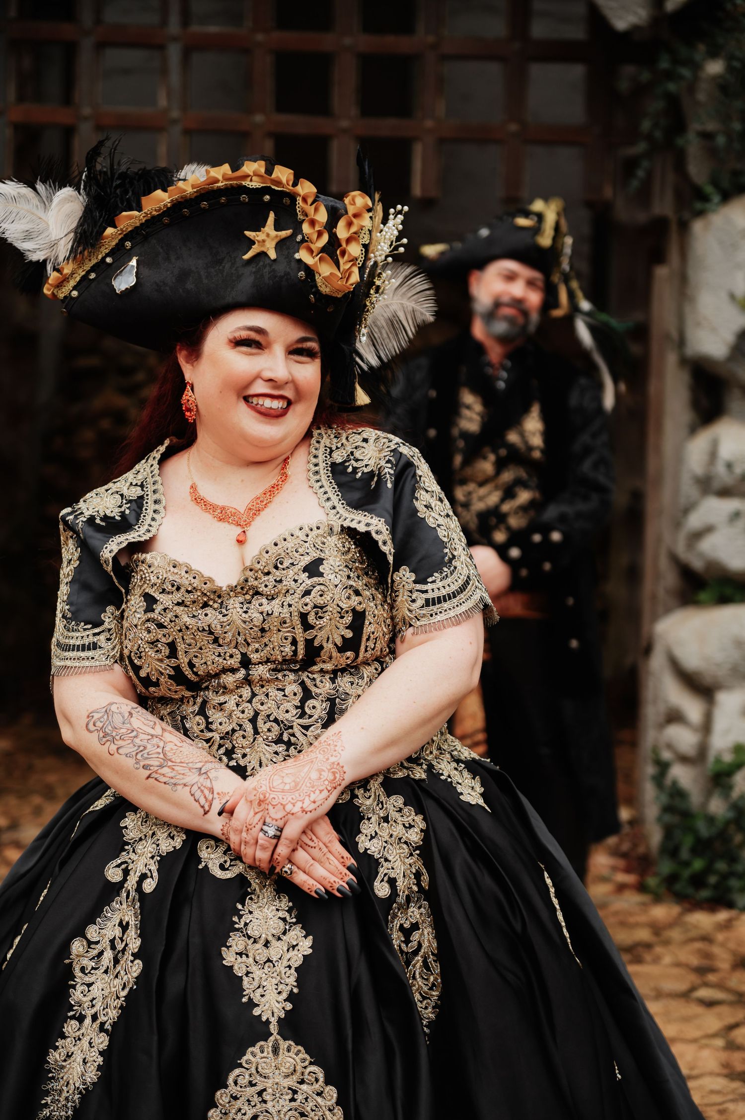 Woman in black gold pirate wedding gown and feathered tricorn hat smiles, man in pirate costume stands behind her.