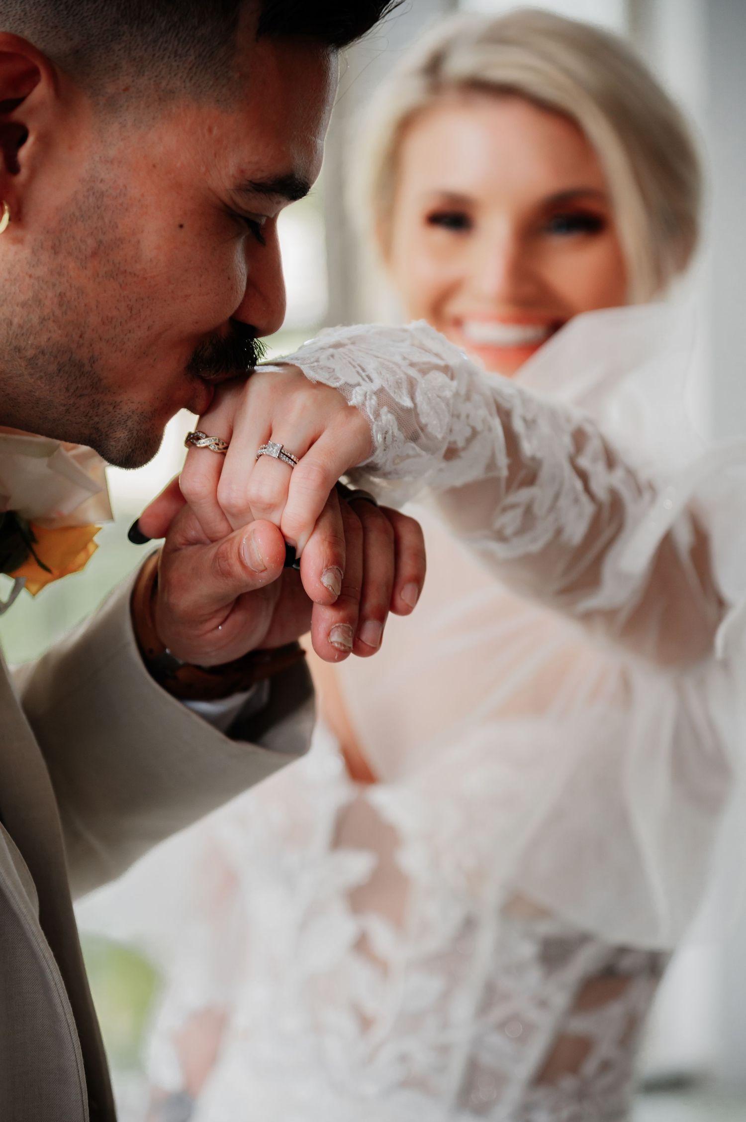 Groom kissing bride's hand showing wedding rings, bride smiling in lace dress and veil in romantic wedding portrait.
