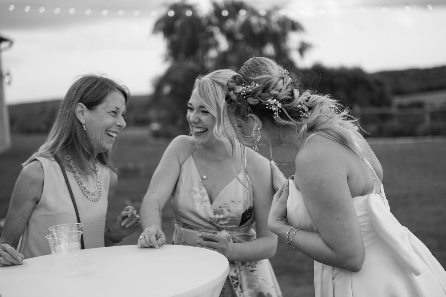 Black and white photo of three women laughing together at a wedding reception outdoor event.