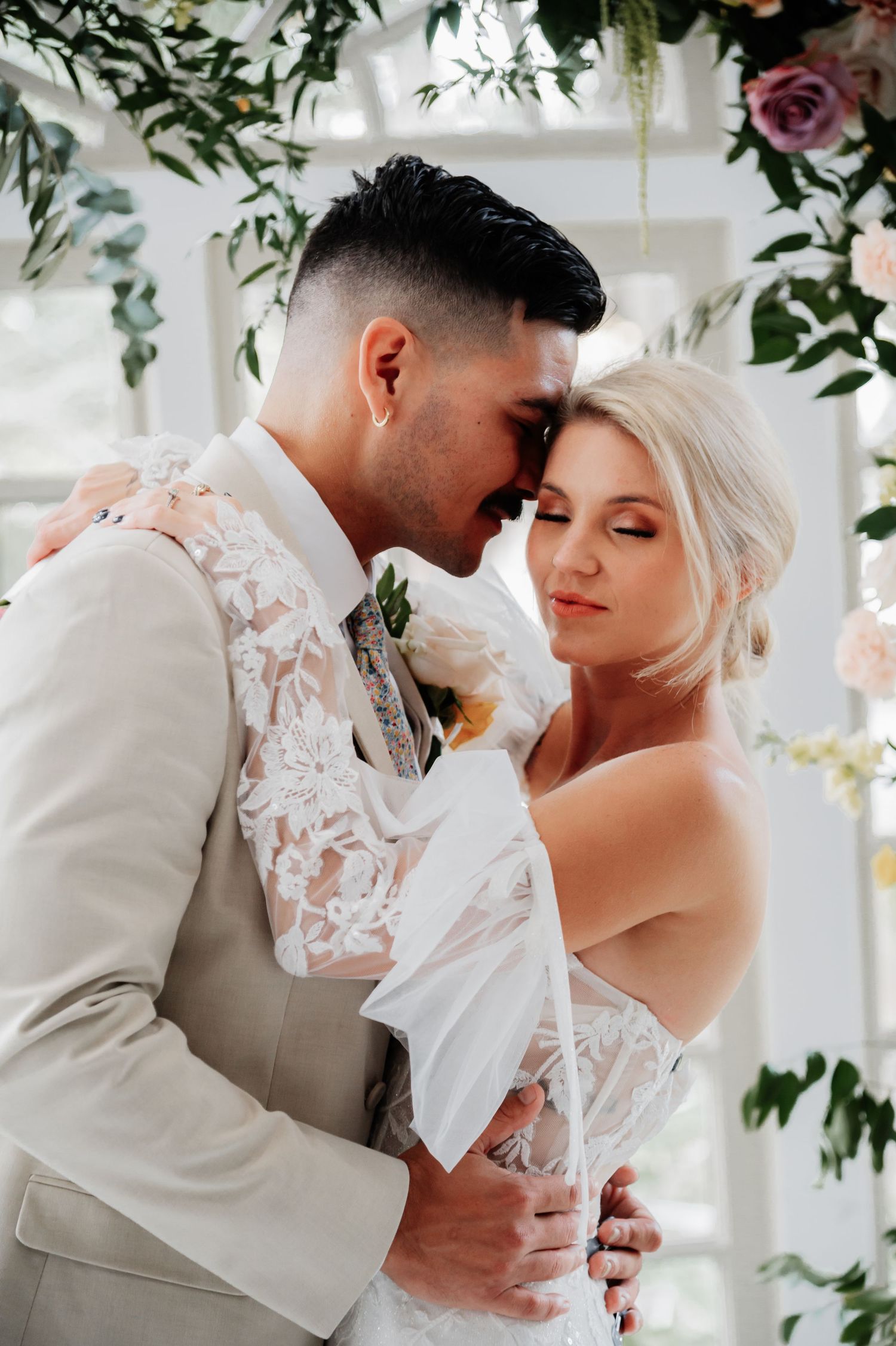 Romantic wedding couple embracing under floral arch, groom in beige suit with lace detail, bride in strapless gown.