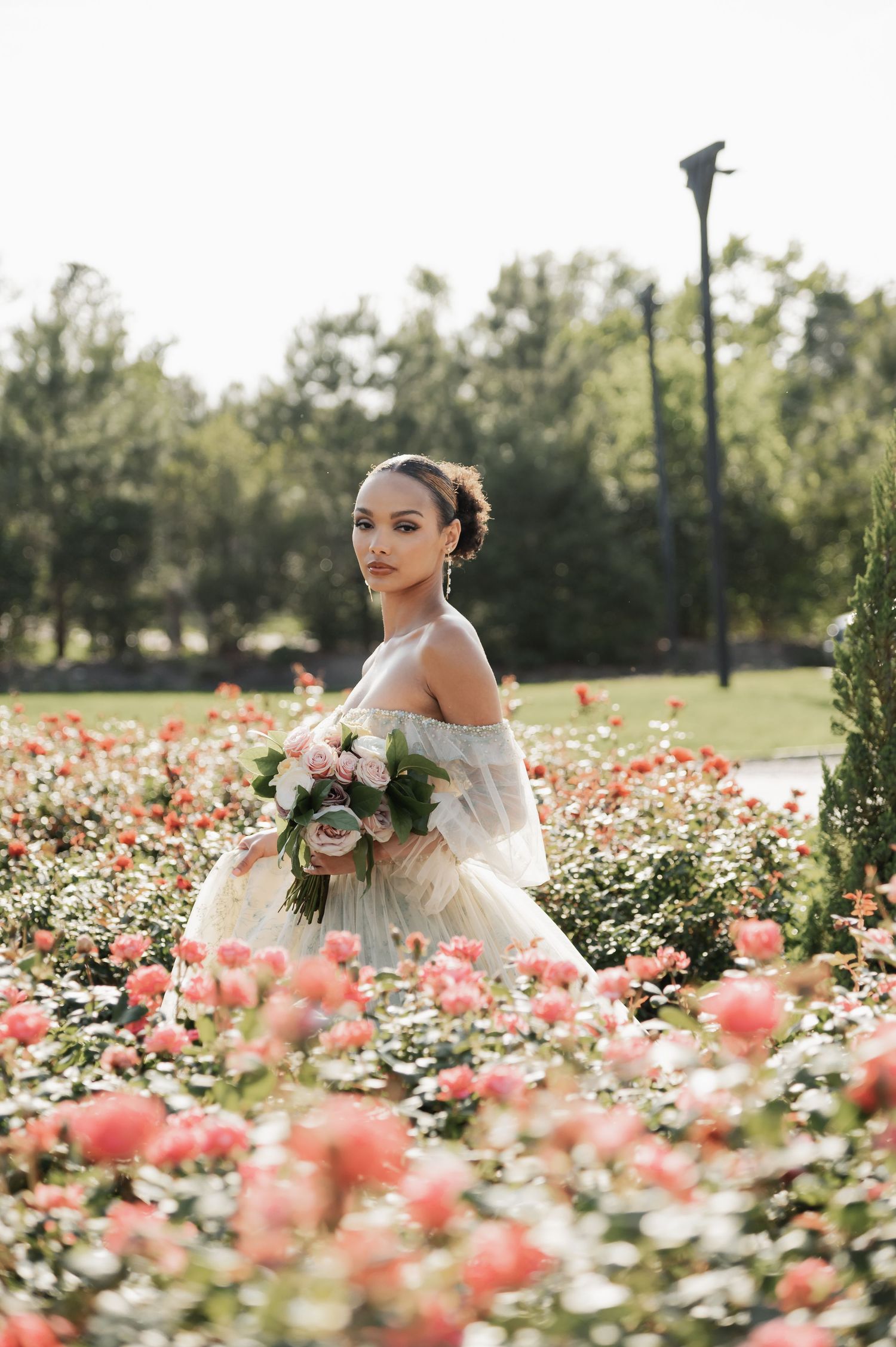 Bride in off-shoulder gown holds bouquet among pink roses in a lush garden setting.