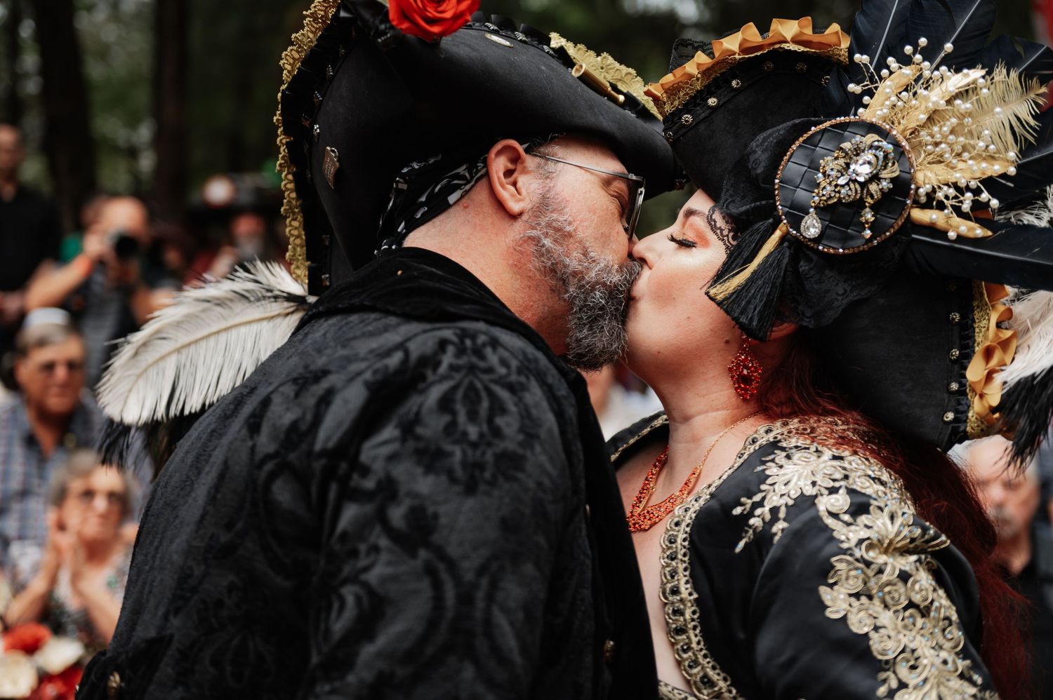 Couple in elaborate pirate costumes sharing a kiss at a masquerade festival outdoors.
