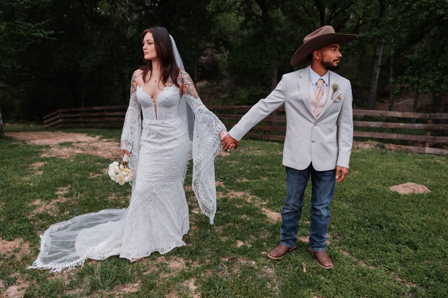Bride in lace gown and groom in cowboy hat hold hands outdoors at rustic wedding venue.