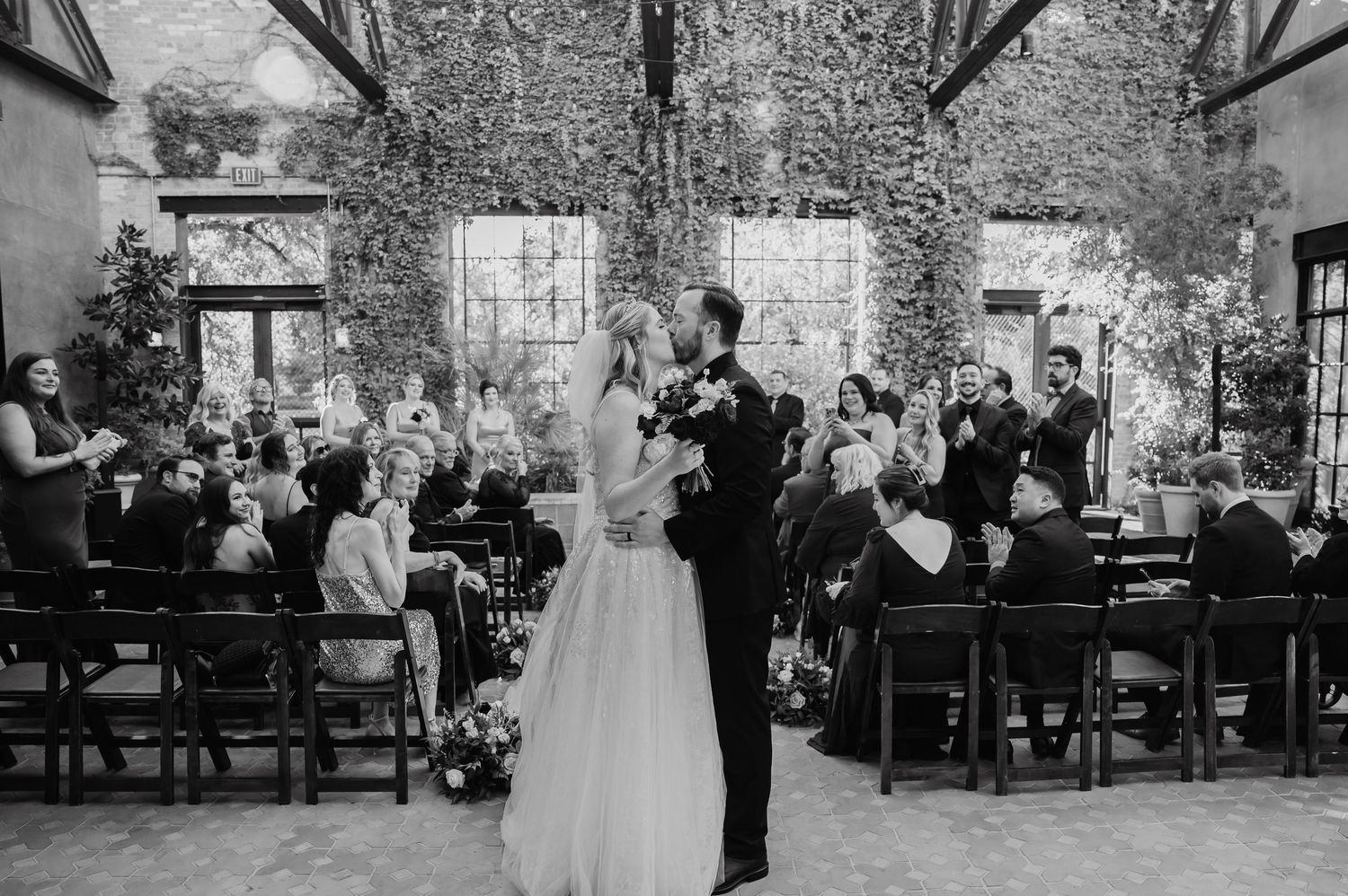 Black and white photo of a bride and groom sharing their first kiss at an elegant ivy-covered wedding venue.