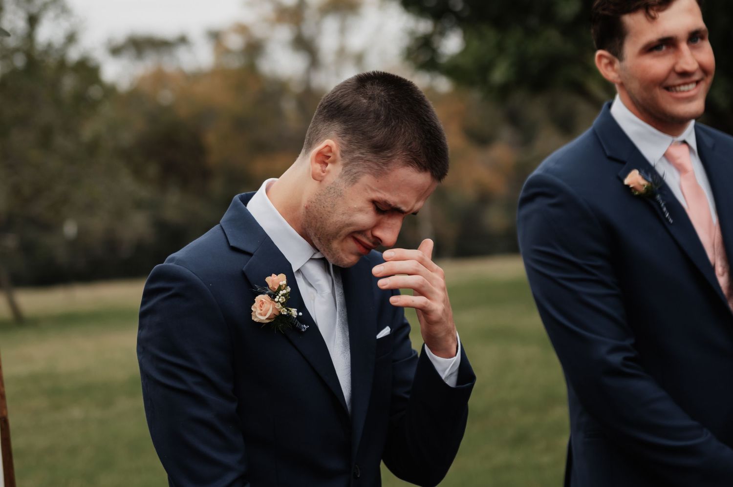 Emotional groom in navy suit crying happy tears at outdoor wedding ceremony with groomsman smiling behind him.