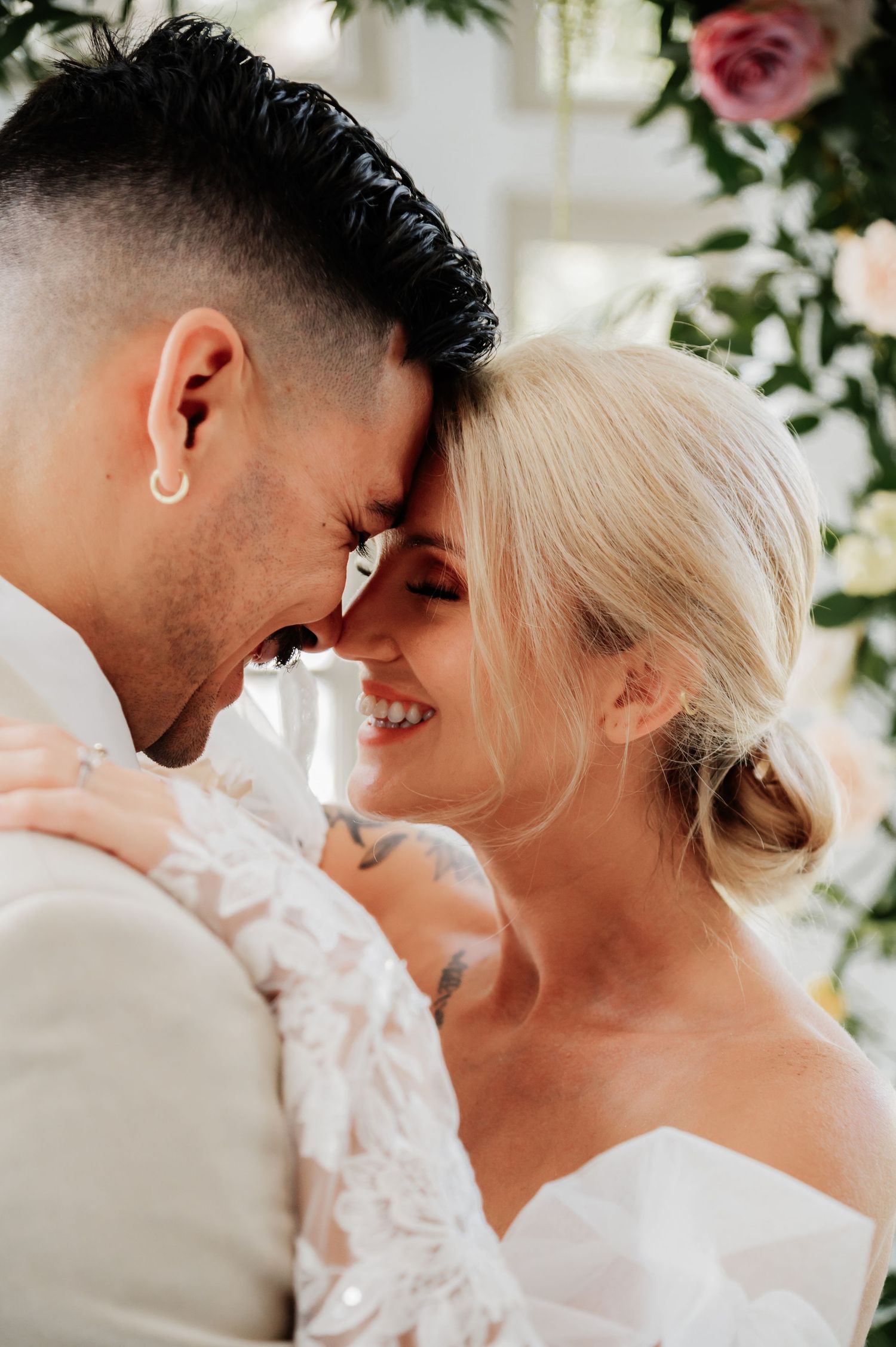 A smiling bride and groom touch foreheads tenderly during their wedding ceremony, surrounded by pink floral decorations.